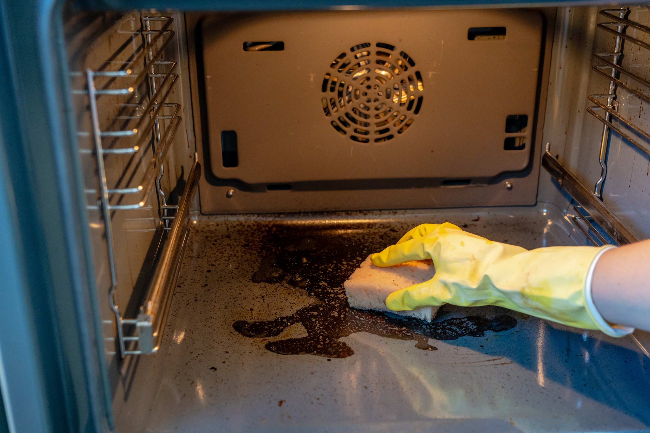 Person wearing yellow rubber gloves cleaning a dirty oven with a sponge, with visible grime and burnt residue inside the oven. The inside of an oven is normally an add on service but in a moving clean it is included.