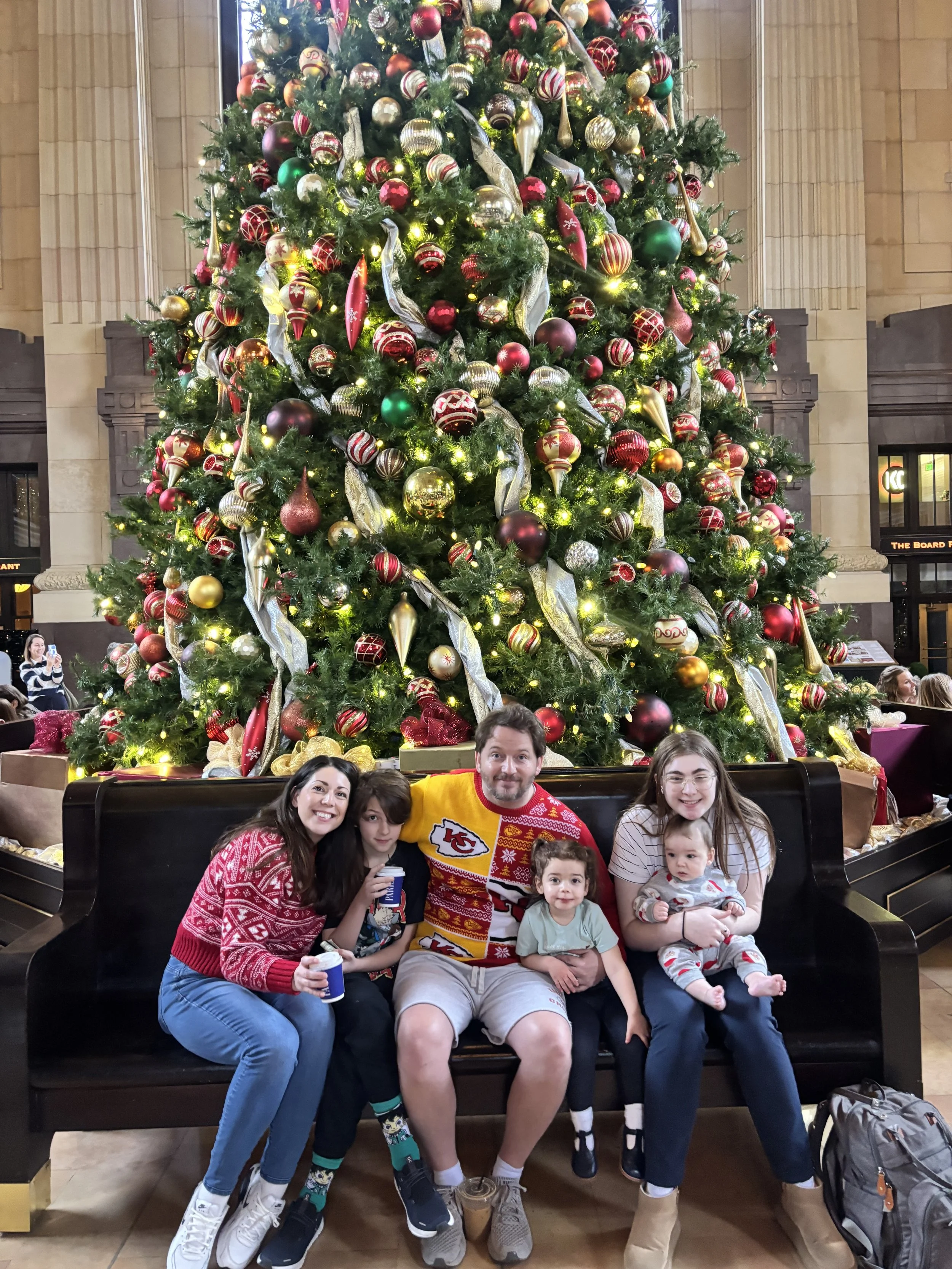 Family of six sitting on a black bench in front of a large decorated Christmas tree, inside a grand hall. The family members are smiling, with Christmas stockings, ornaments, and lights on the tree.
