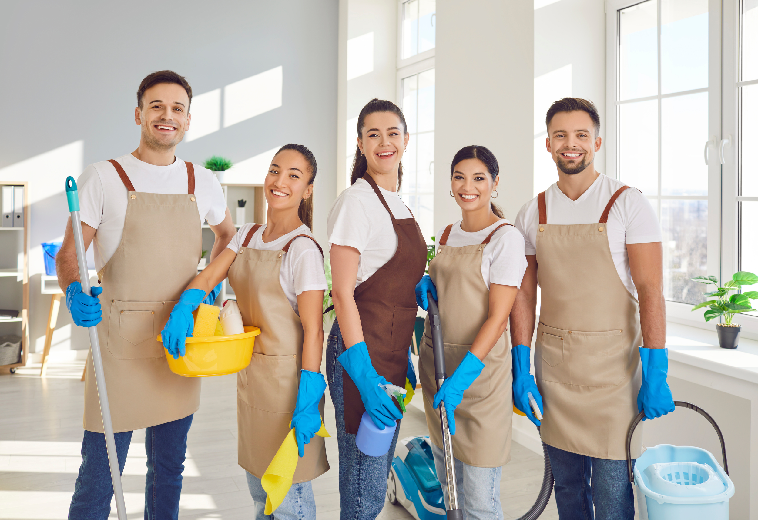 Group of five diverse cleaning staff members smiling and posing with cleaning tools in a bright, modern room with large windows and indoor plants.