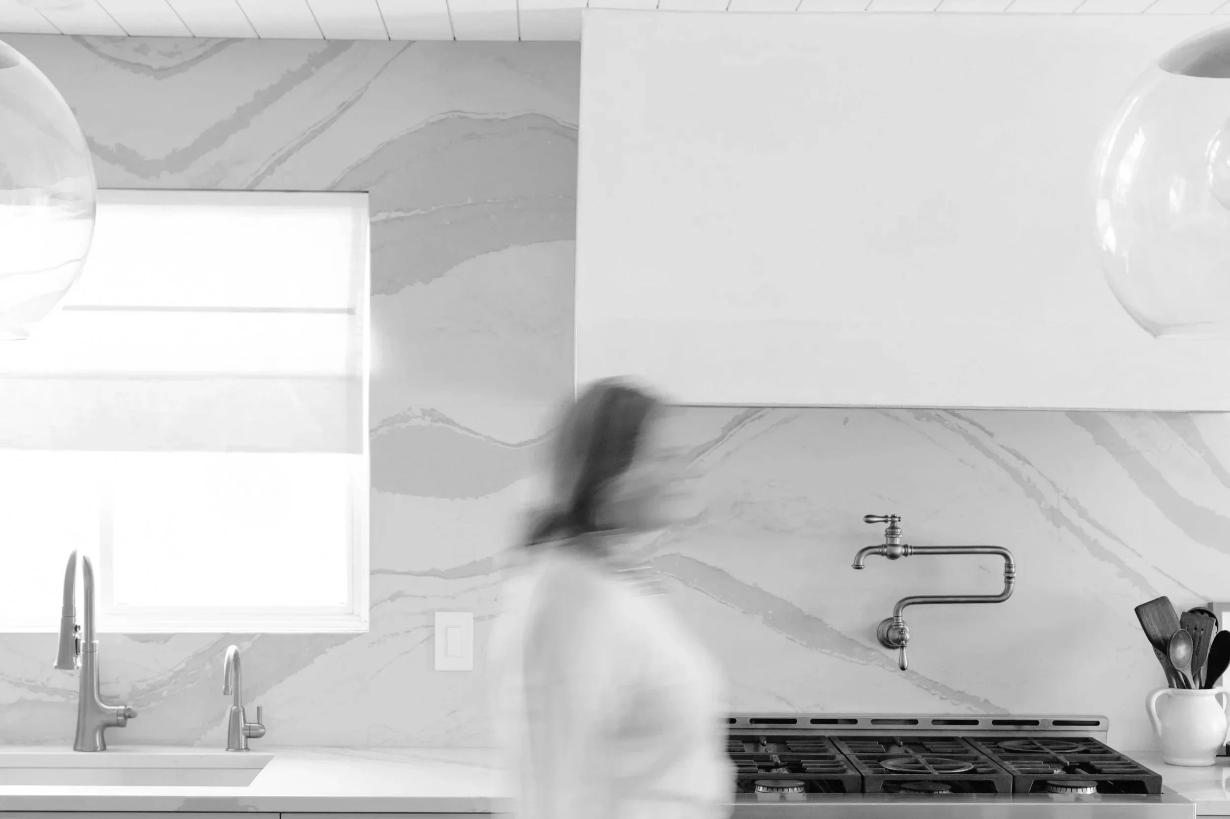 A modern kitchen with a marble backsplash, a window, a double sink with a faucet, a stovetop, a wall-mounted faucet, a white mug with kitchen utensils, and a blurred person walking through.