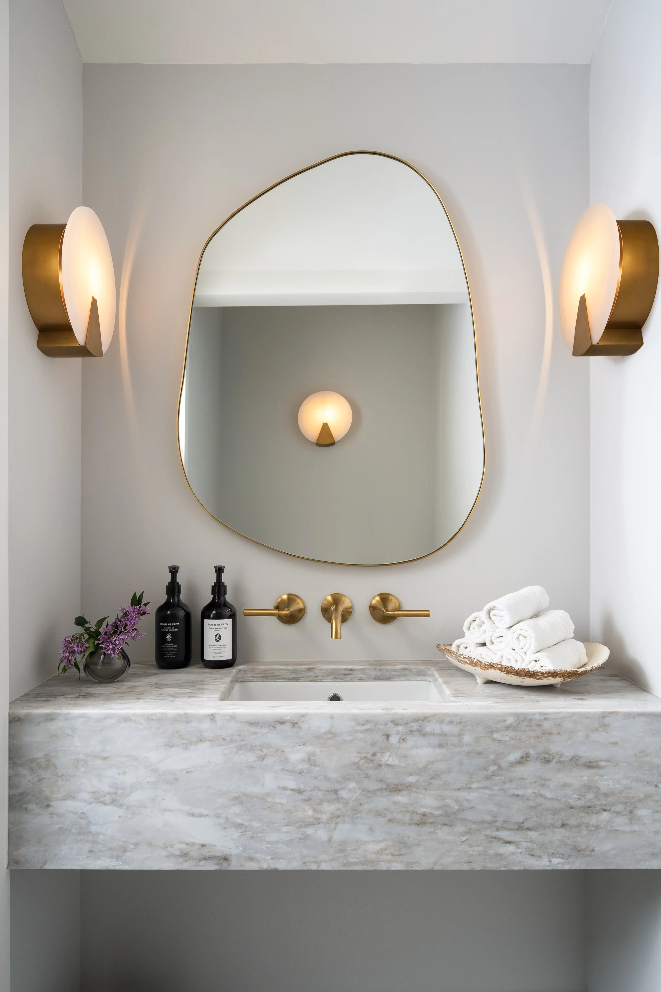 Modern bathroom vanity with a marble countertop, a large gold-framed mirror, two gold wall sconces, a flower vase with purple flowers, two soap dispensers, a brass faucet, and a rolled towel on a plate.