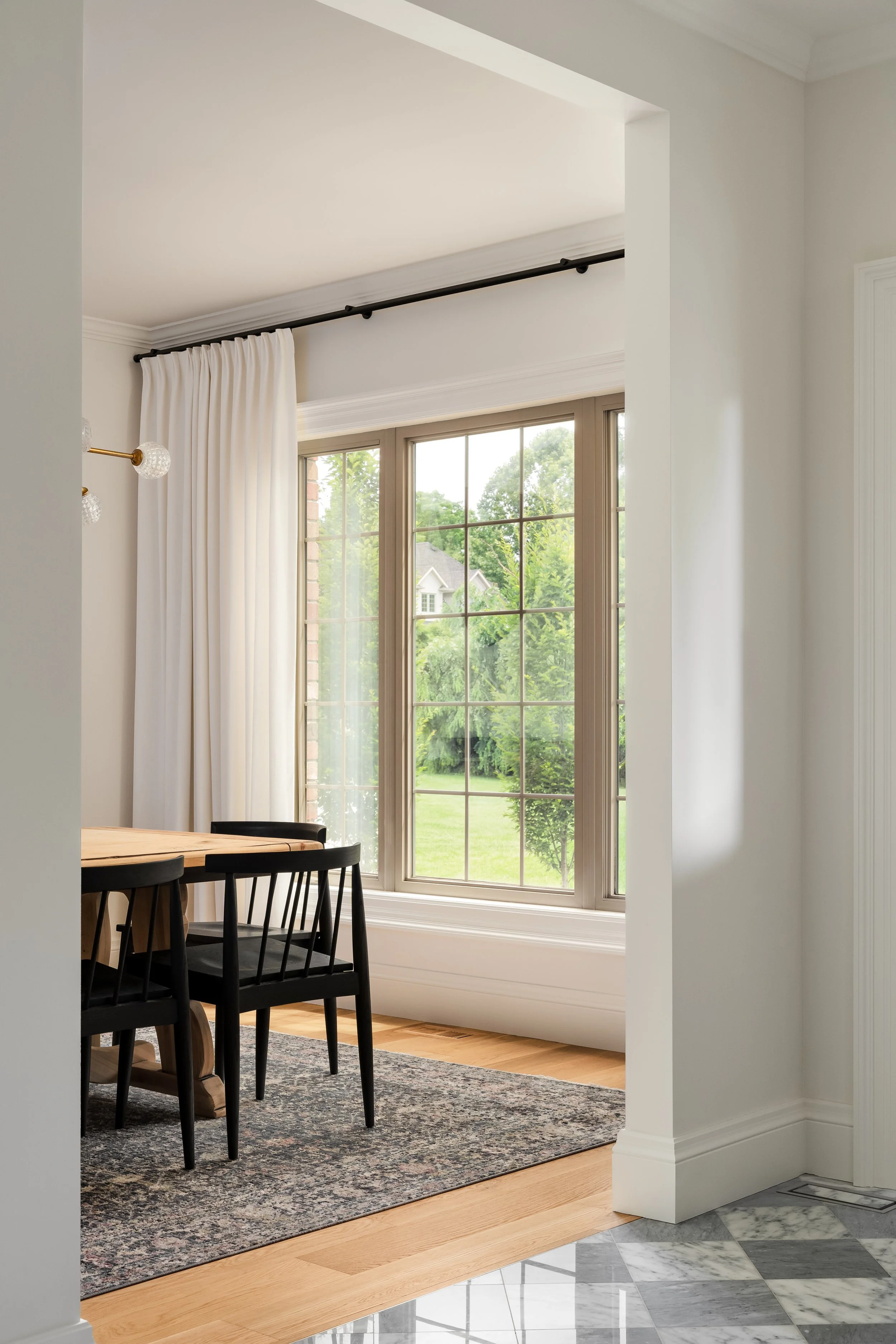 View of a dining area with a large window, white curtains, black chairs, a wooden table, and a patterned area rug.