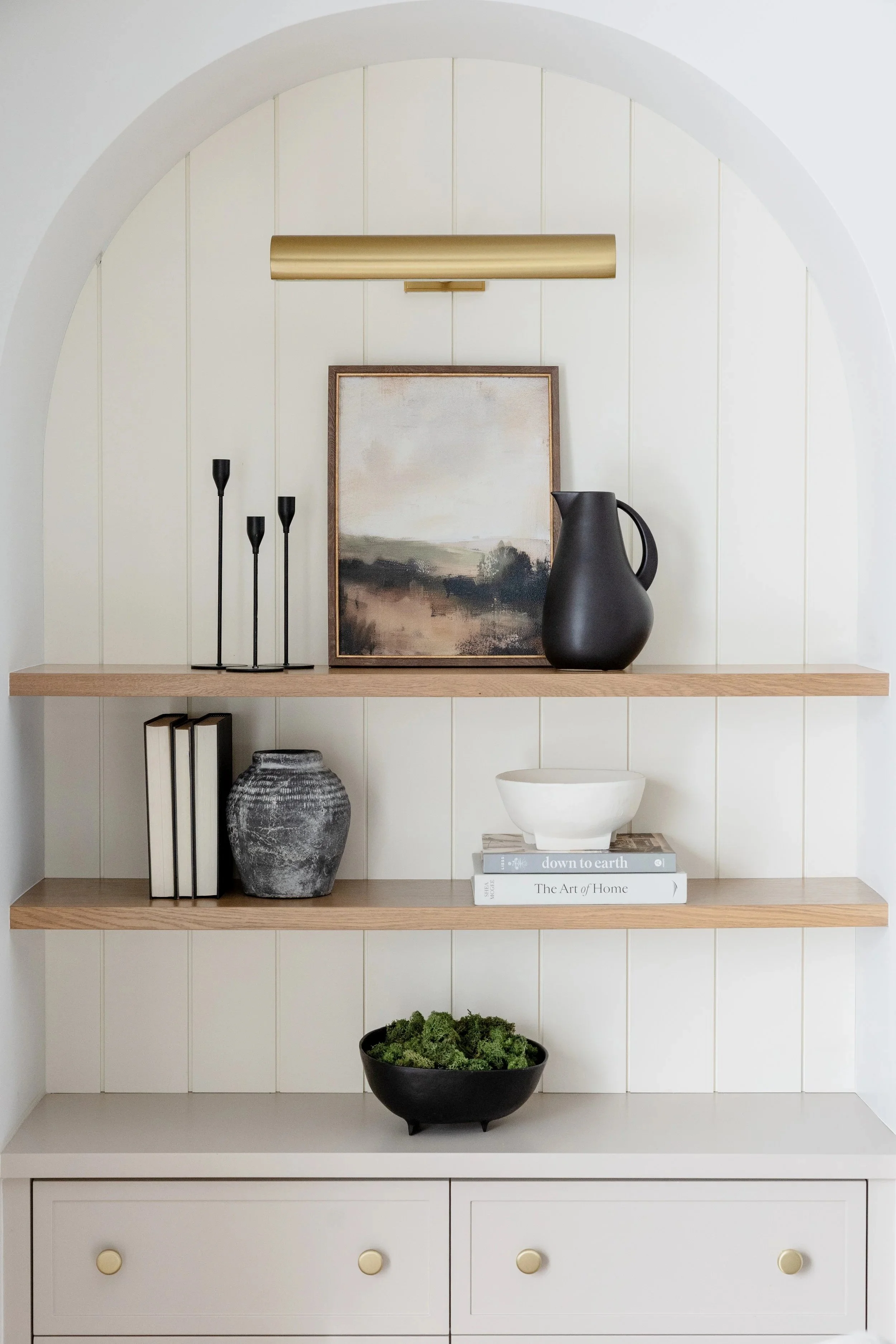 Wooden shelves with decorative items including books, a black pitcher, a gray vase, a bowl with green moss, and artwork, in a white arched alcove.