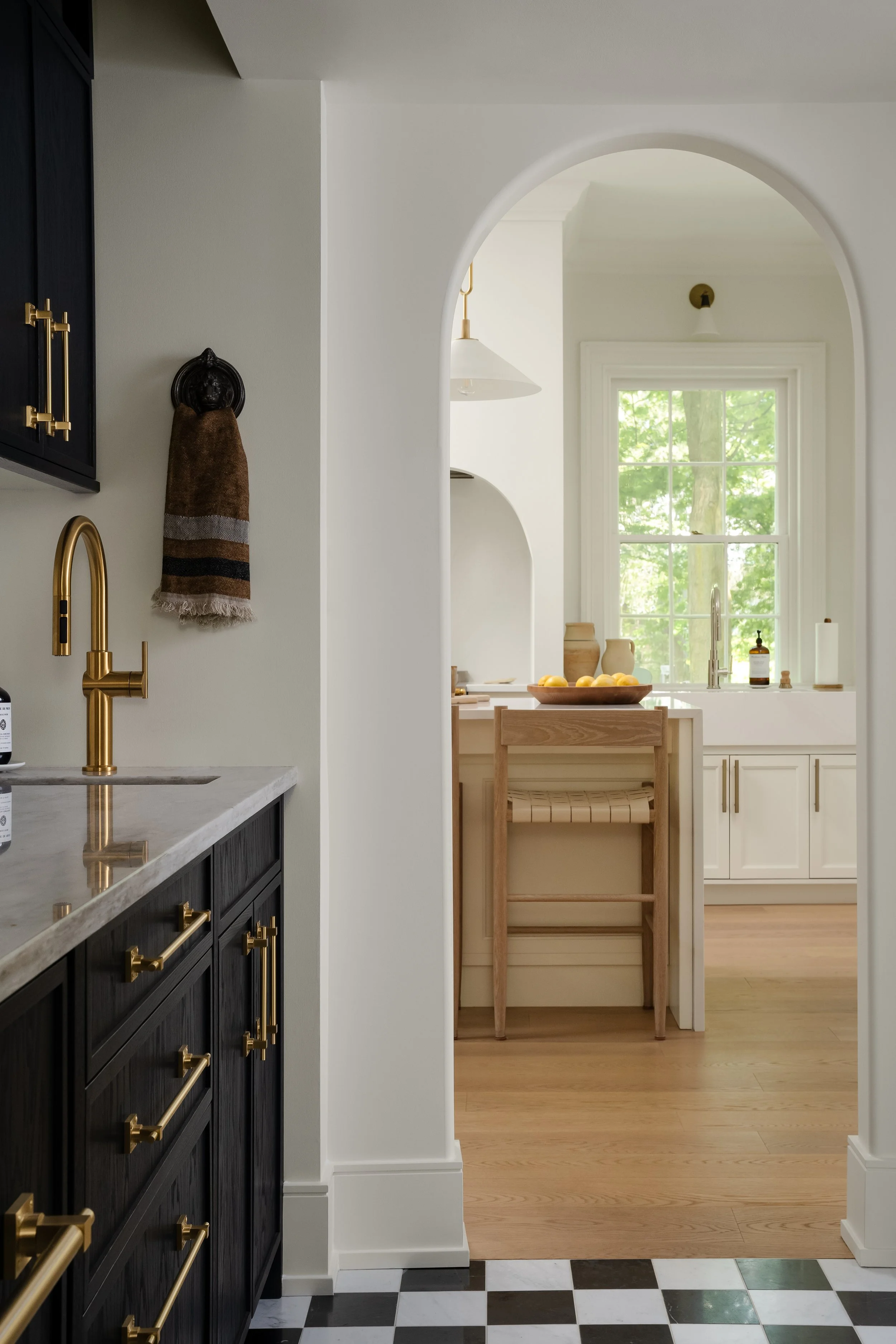 View of a kitchen with black cabinets, gold hardware, a gold faucet, a marble countertop, and a black and white checkered floor, seen through an arched doorway leading to a bright white kitchen with a large window and light wood flooring.