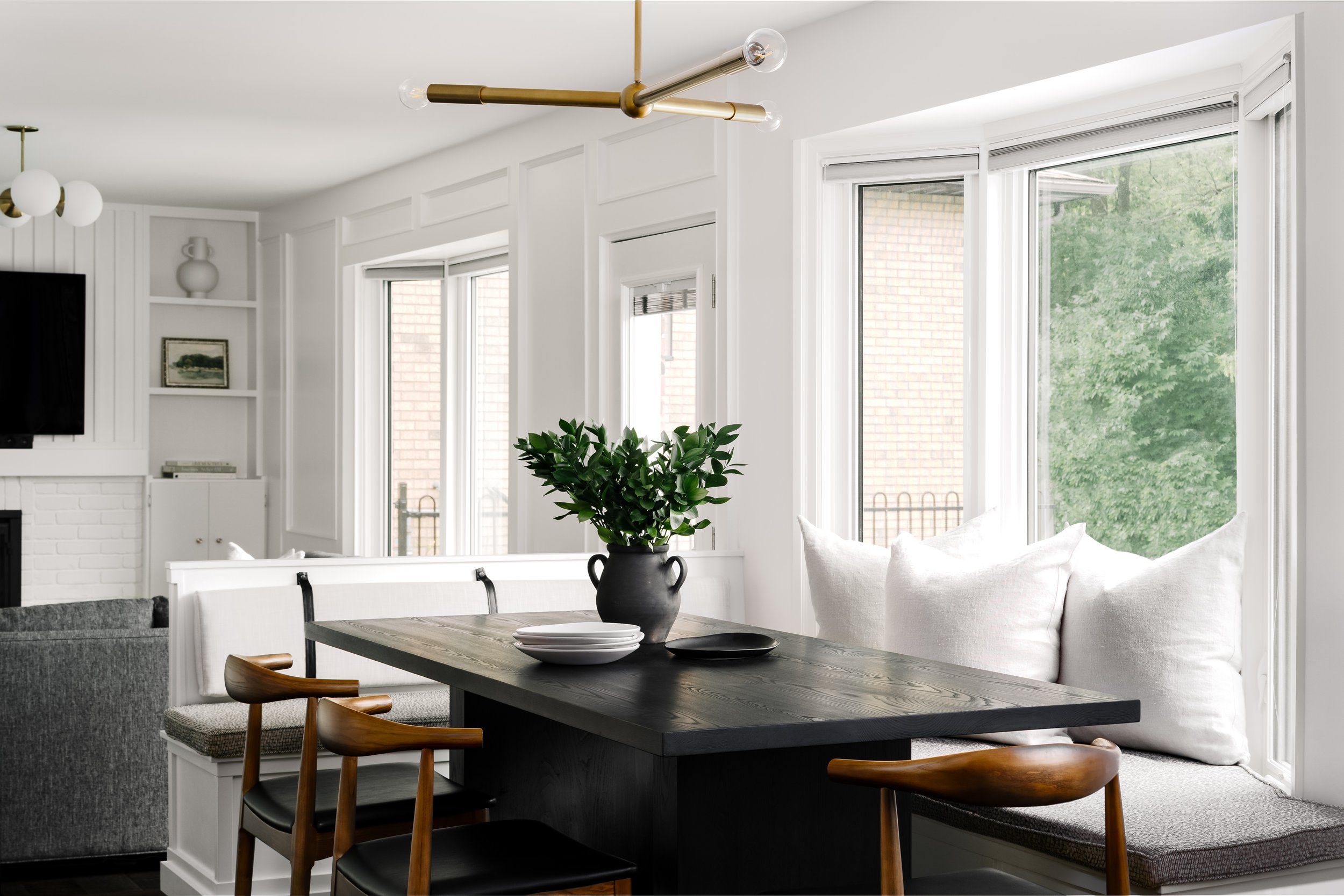Bright dining area with large windows, a black table, wooden chairs, white cushions, and a green plant centerpiece.
