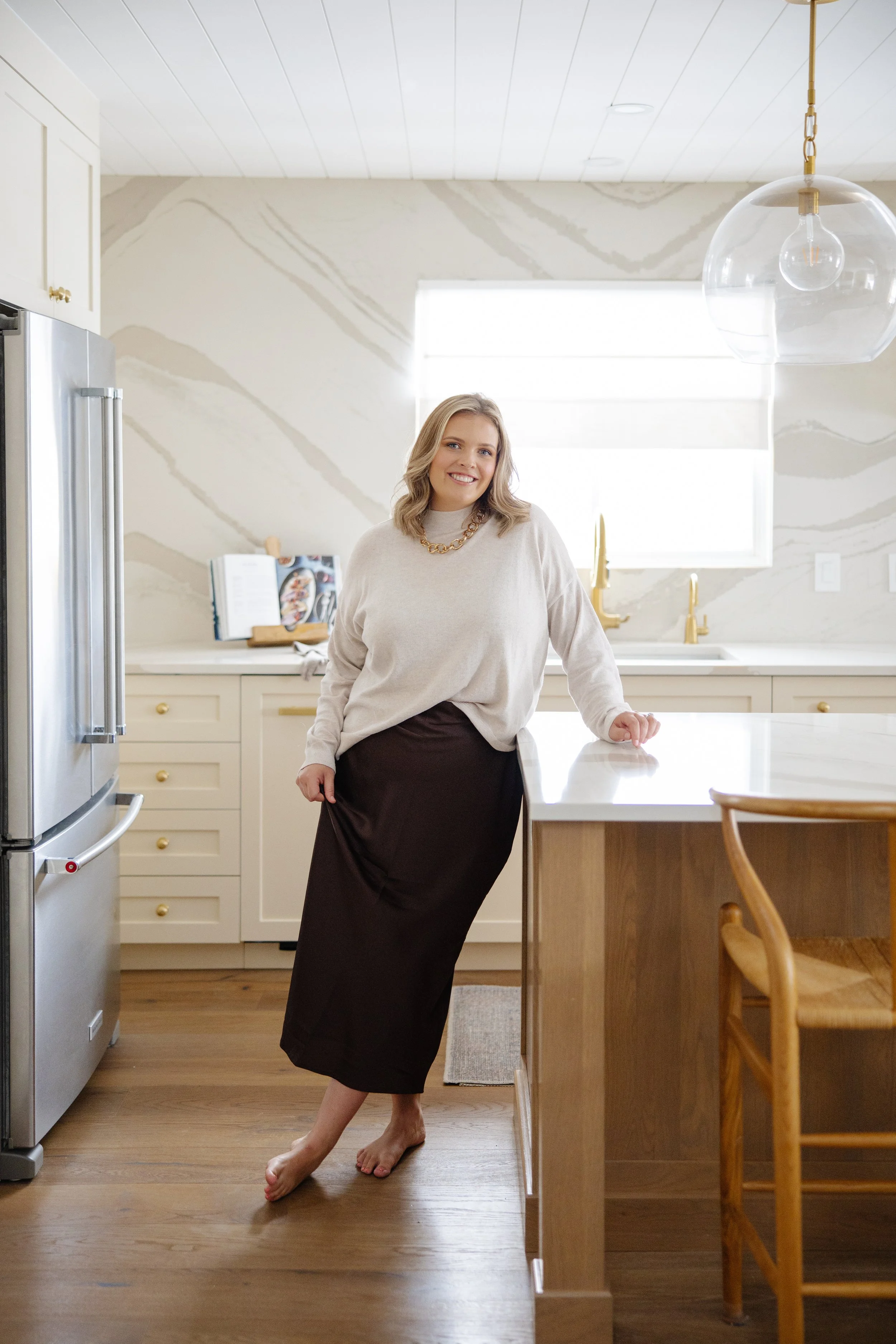 A woman with blonde hair wearing a beige sweater and a black skirt standing barefoot in a modern kitchen, smiling at the camera.