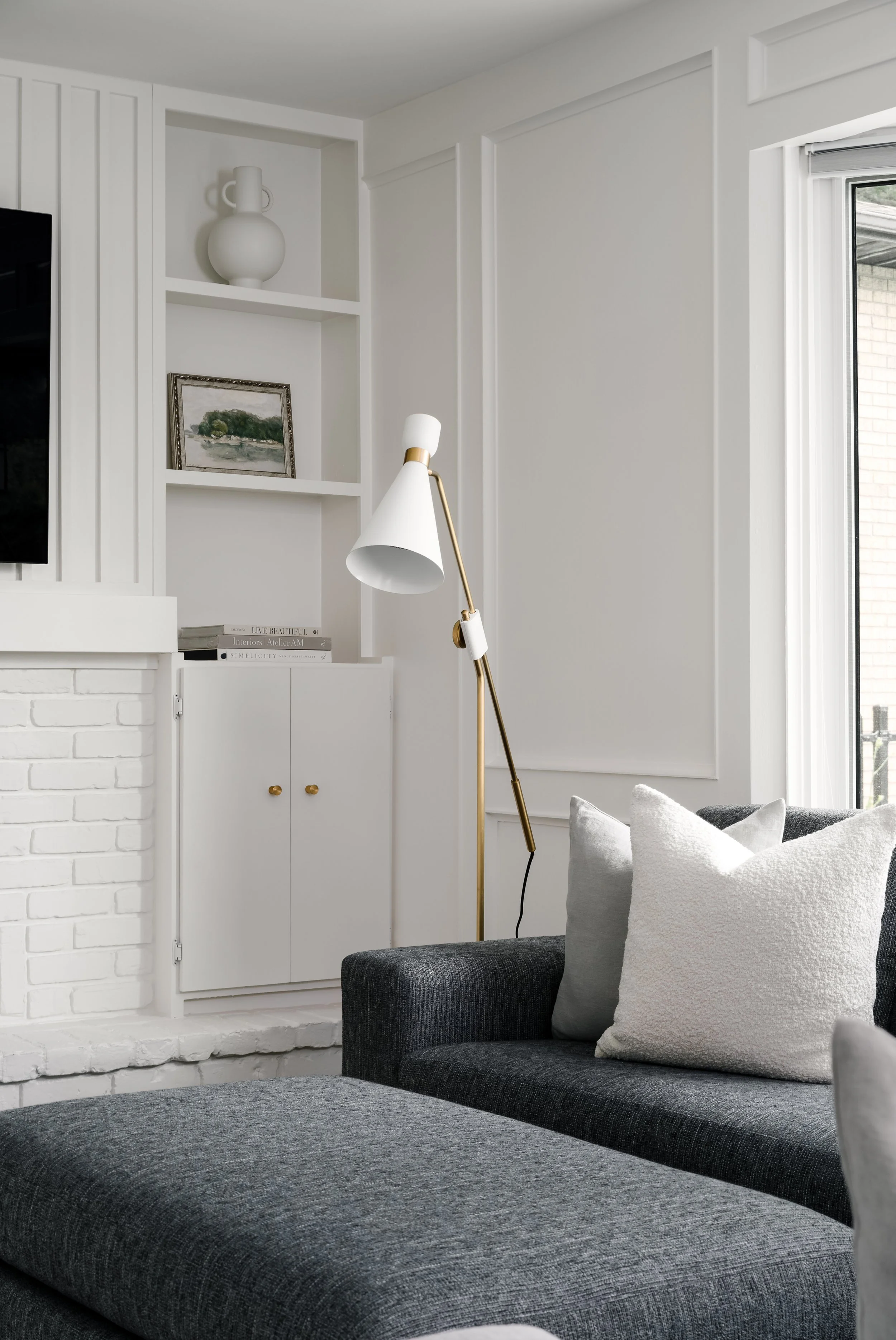 A cozy living room corner featuring a dark gray sofa with light gray and white pillows, a white wall with decorative paneling, a white bookshelf with a painting, a ceramic vase, and a white and gold floor lamp near a window.