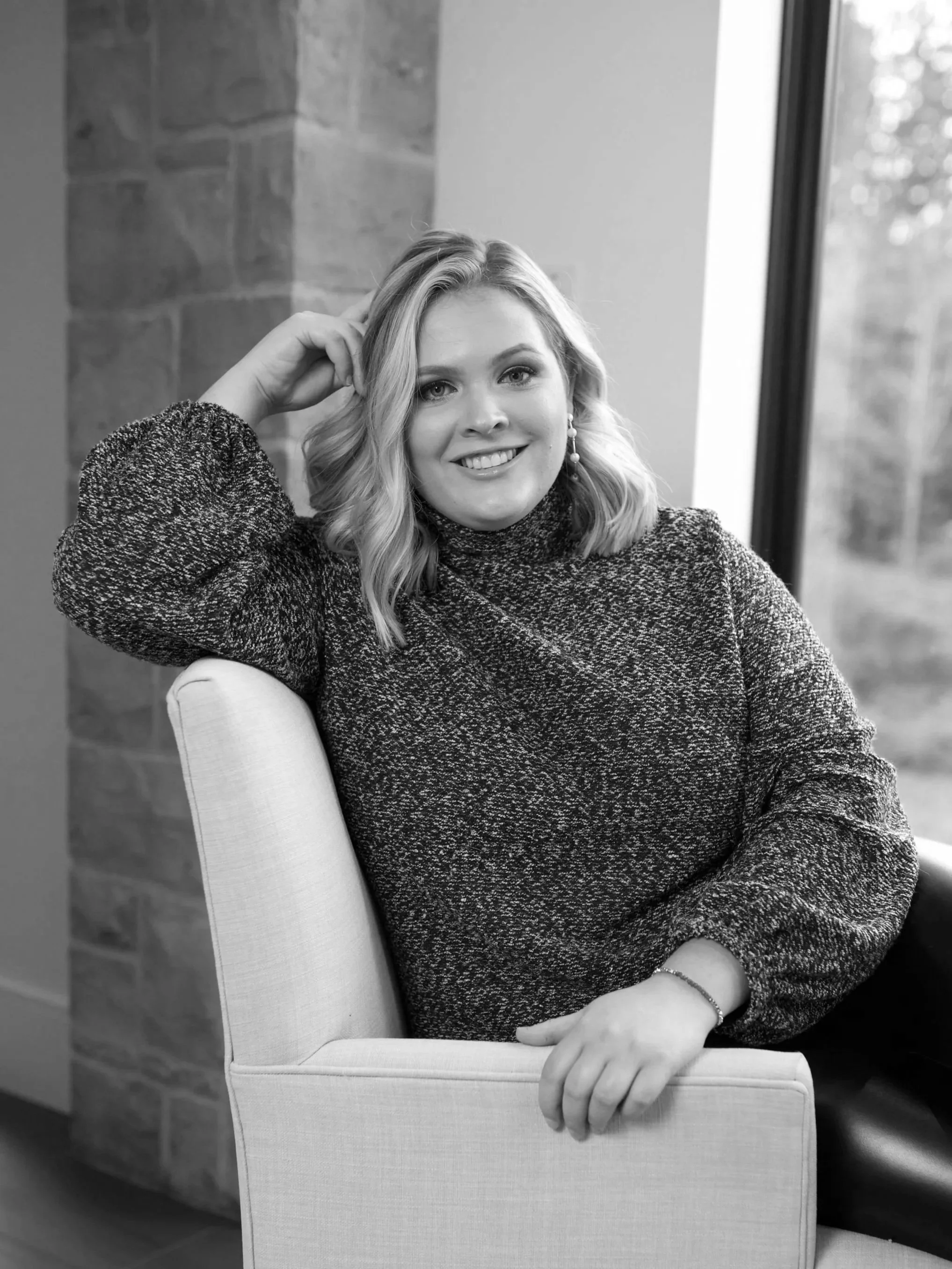 Black and white photo of a young woman with wavy blonde hair sitting on a chair near a window, smiling at the camera, wearing a textured long-sleeve top and earrings.