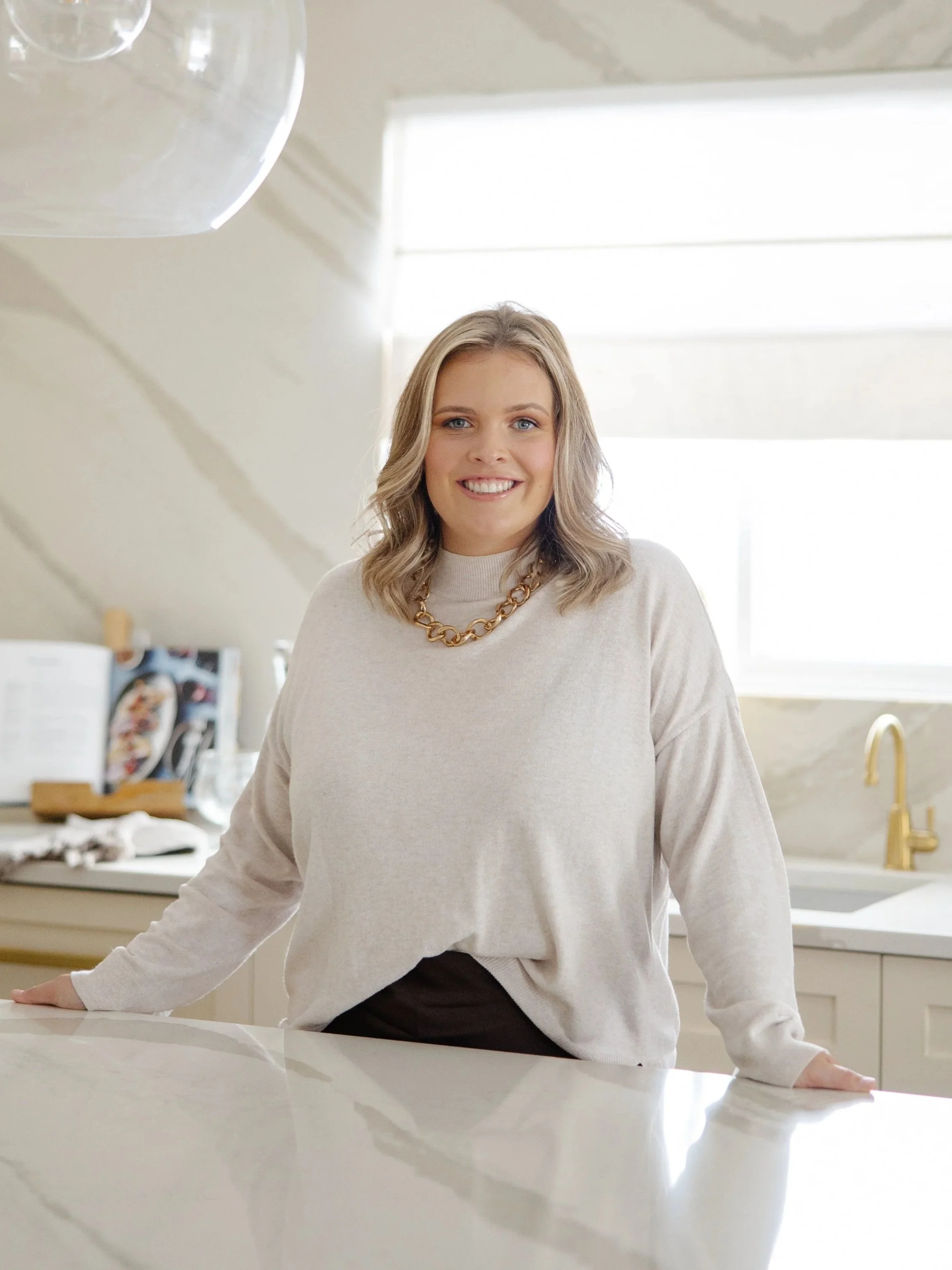 A woman with blonde hair smiling and leaning on a kitchen counter with a bright, well-lit kitchen in the background.