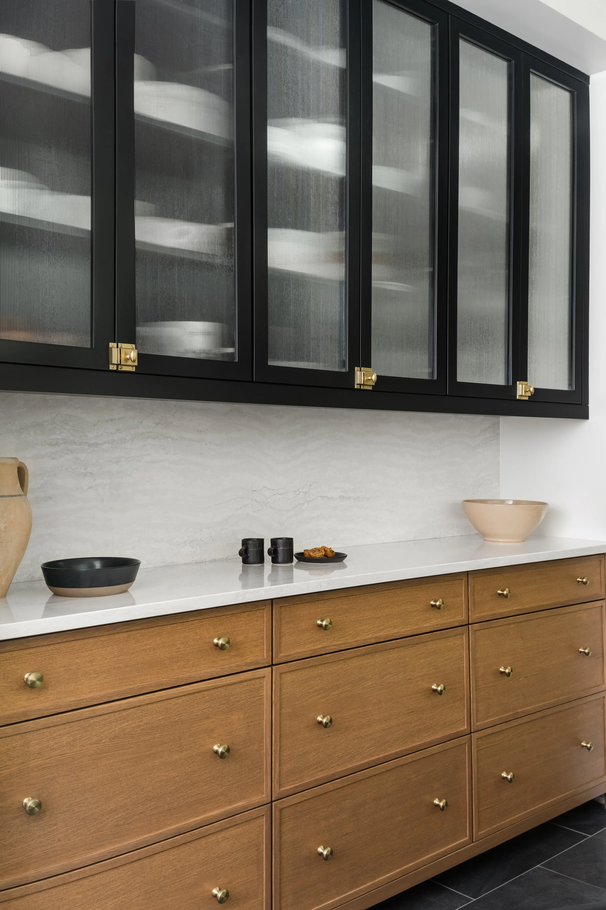 Kitchen cabinet with a black upper glass-fronted cabinet and a wooden lower cabinet with gold knobs, along with some dishes and cups on the countertop.
