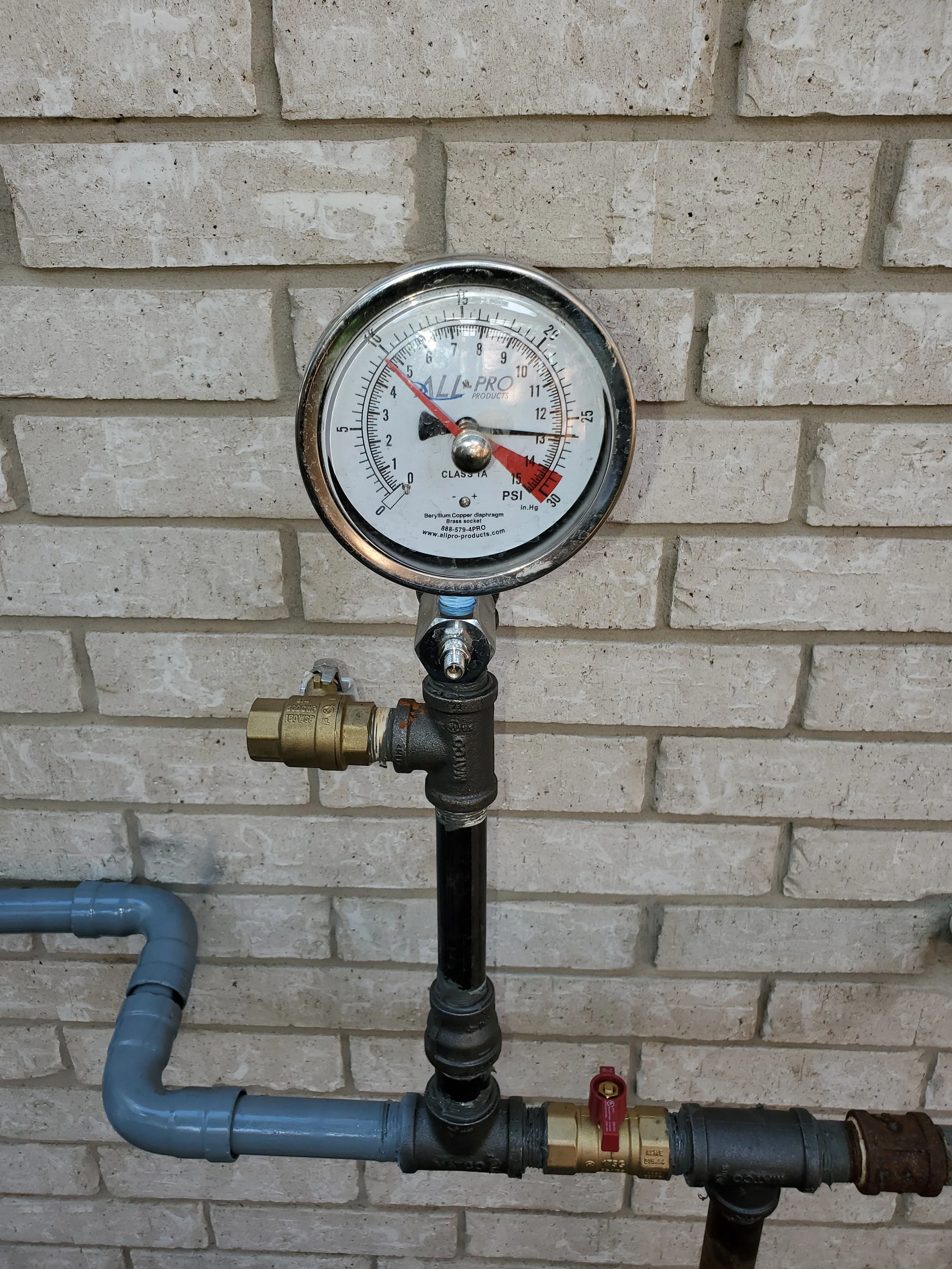 A water pressure gauge attached to plumbing pipes on a brick wall, with a brass valve and a shut-off valve below.