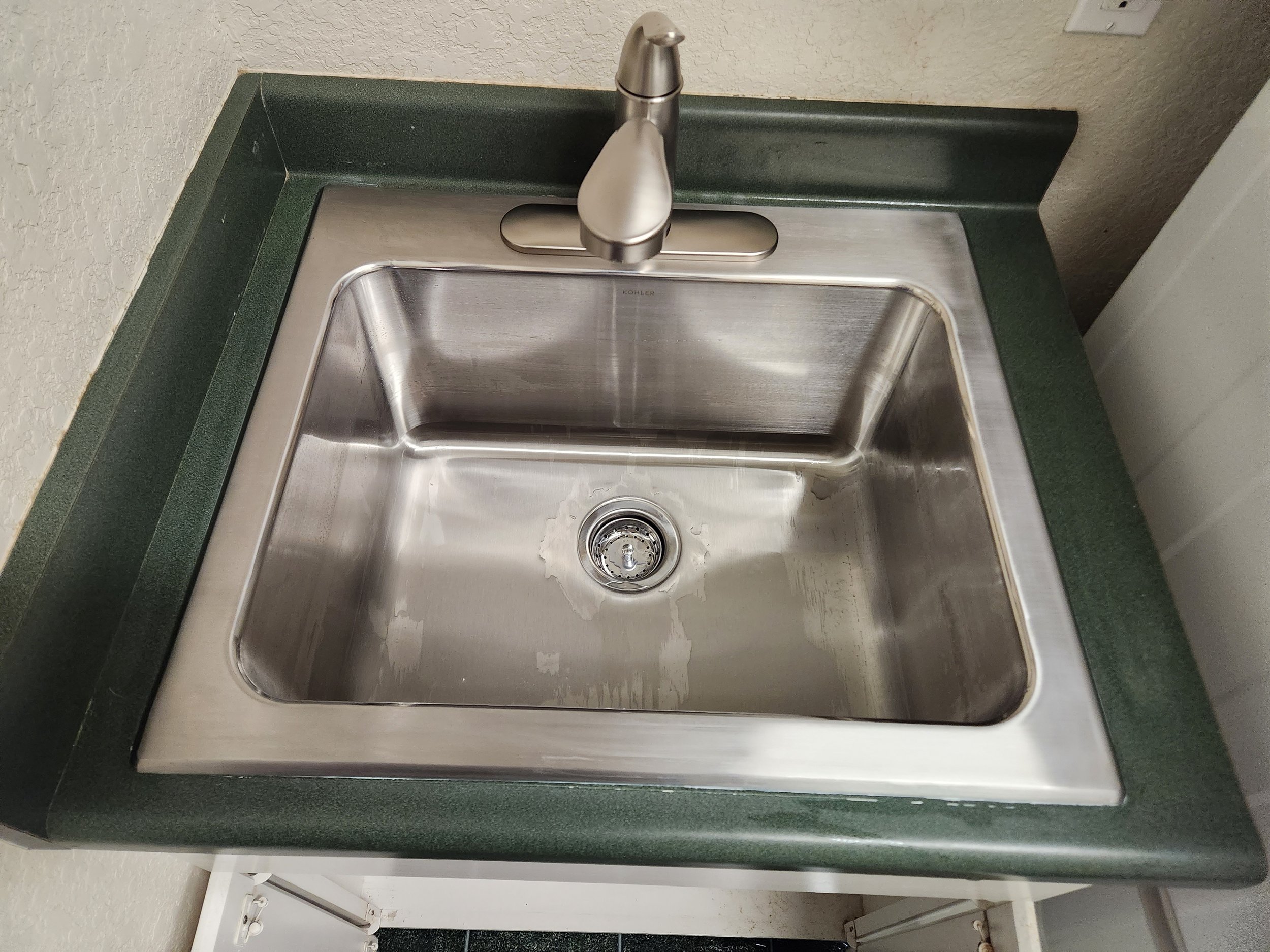 A stainless steel kitchen sink with a green countertop and a brushed nickel faucet, mounted against a textured beige wall.