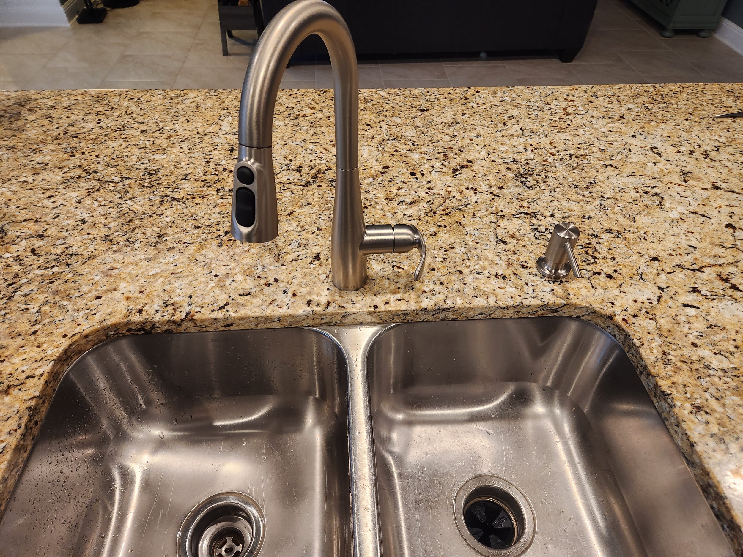 Stainless steel double kitchen sink with granite countertop, modern pull-down faucet, soap dispenser, and a black handle on the faucet.
