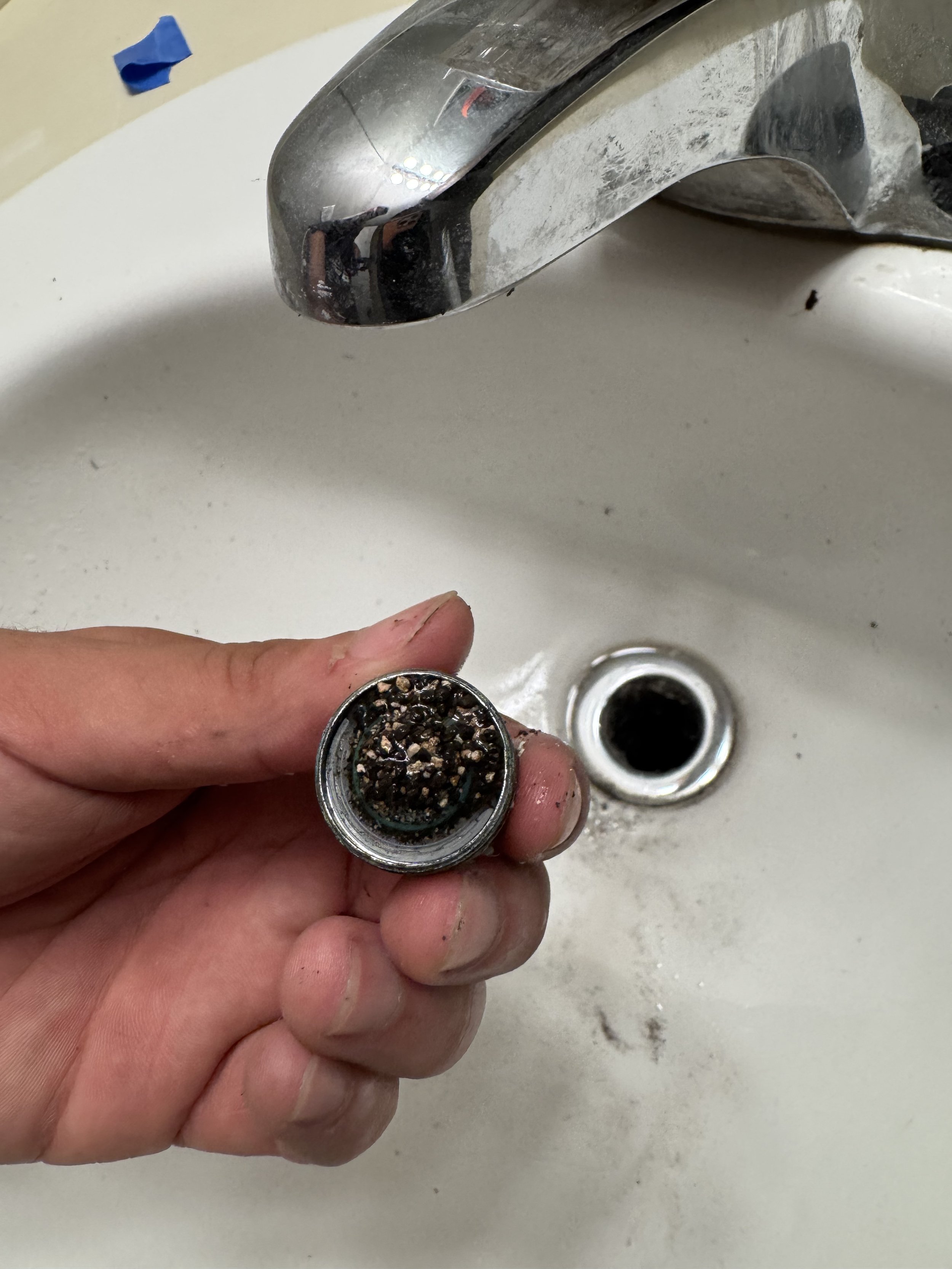 Hand holding a clogged faucet screen filter over a white sink, with a chrome faucet above and a drain below.