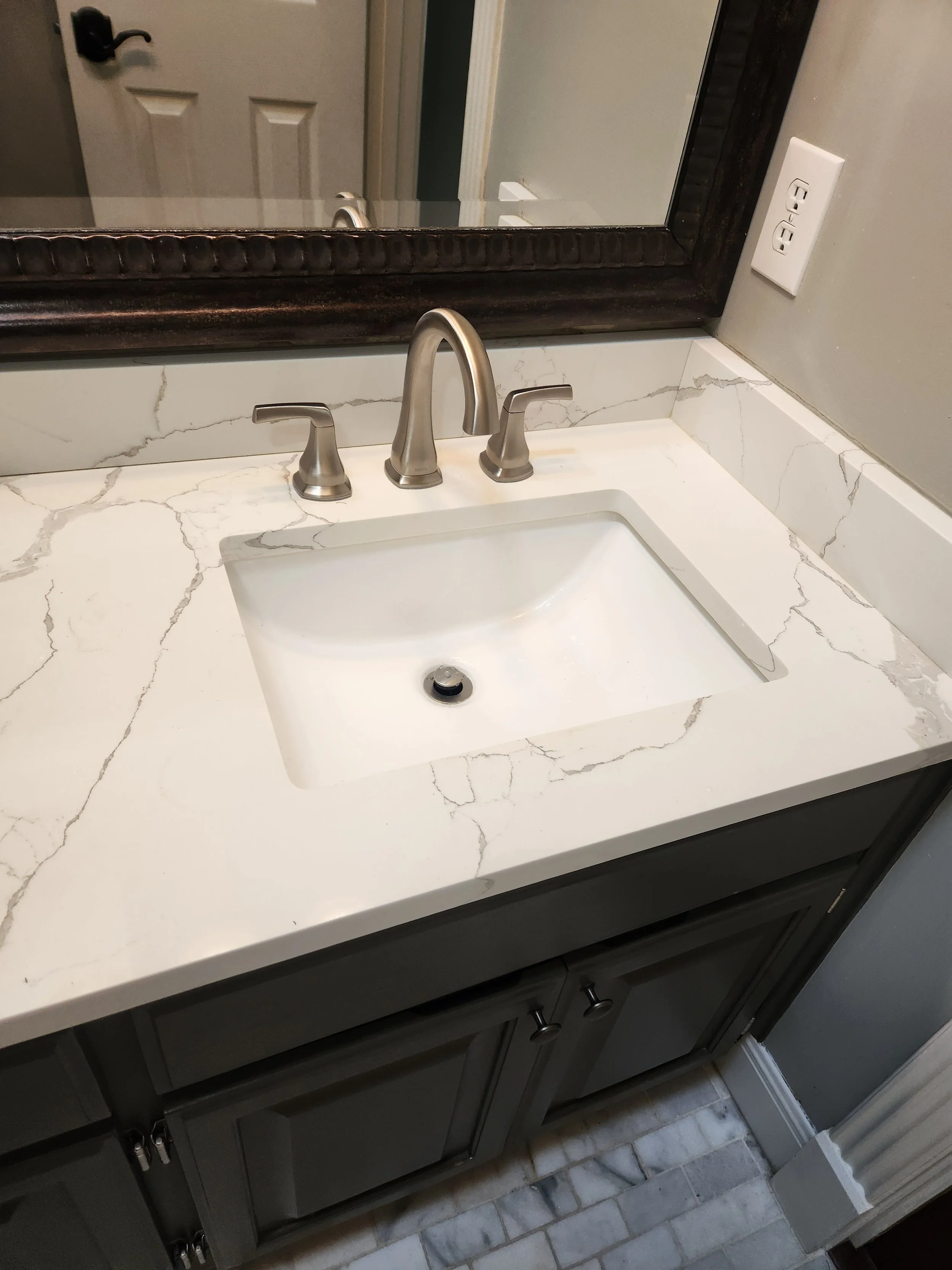 Bathroom vanity with a white marble countertop, an undermount sink, and a brushed nickel faucet with two handles, reflected in a mirror above the vanity.