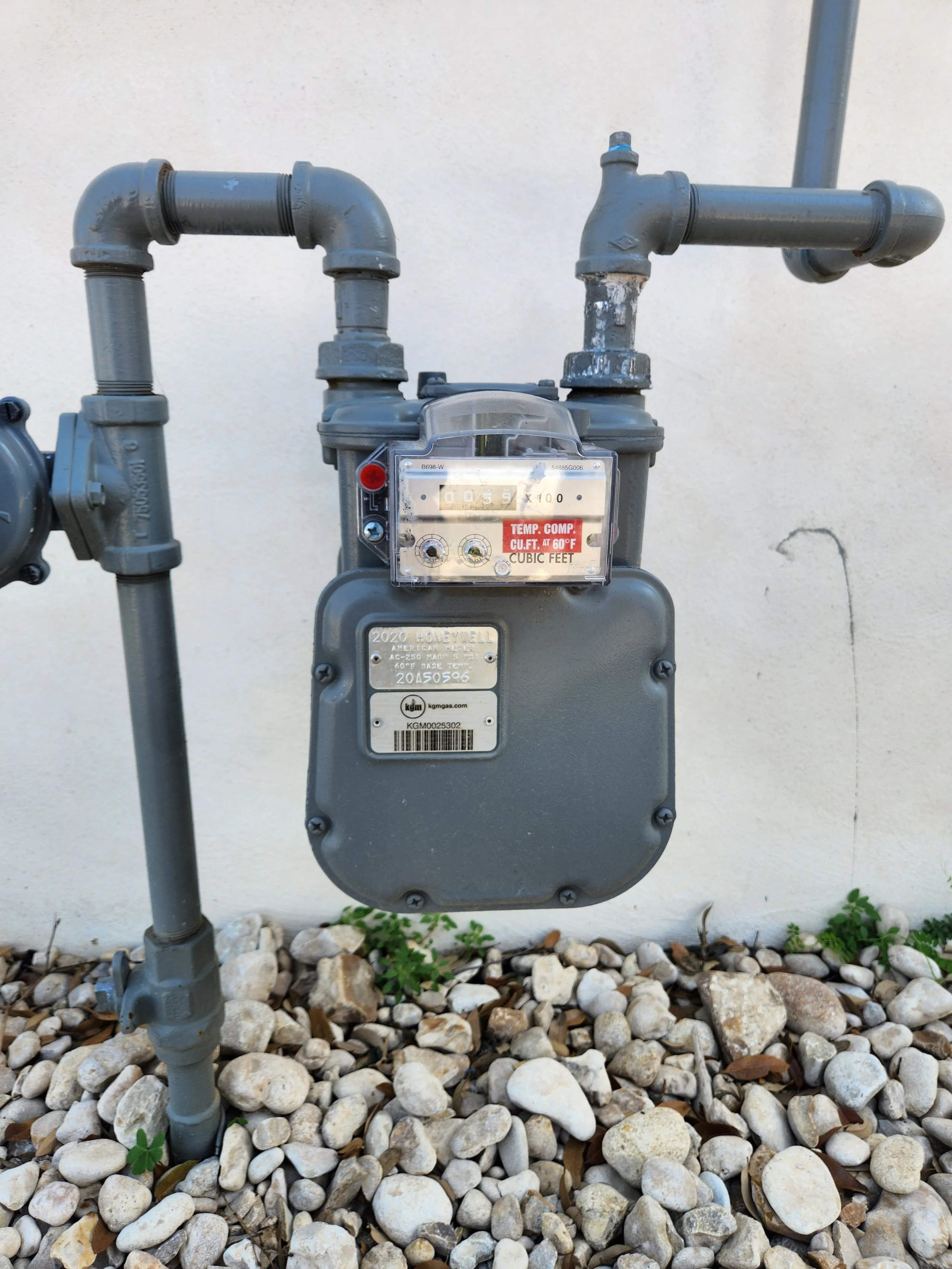 An outdoor natural gas meter attached to a beige wall with small rocks and some green plants at the base.