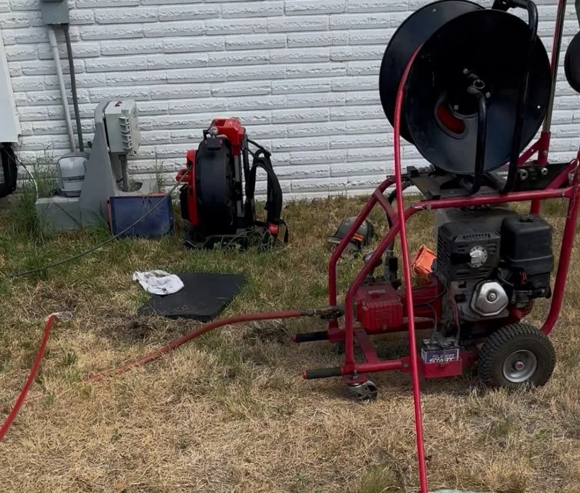 Portable high-pressure water jetting machine with a red frame, large black hose reel, and a small engine, set up on a patch of dry grass near a white brick wall.
