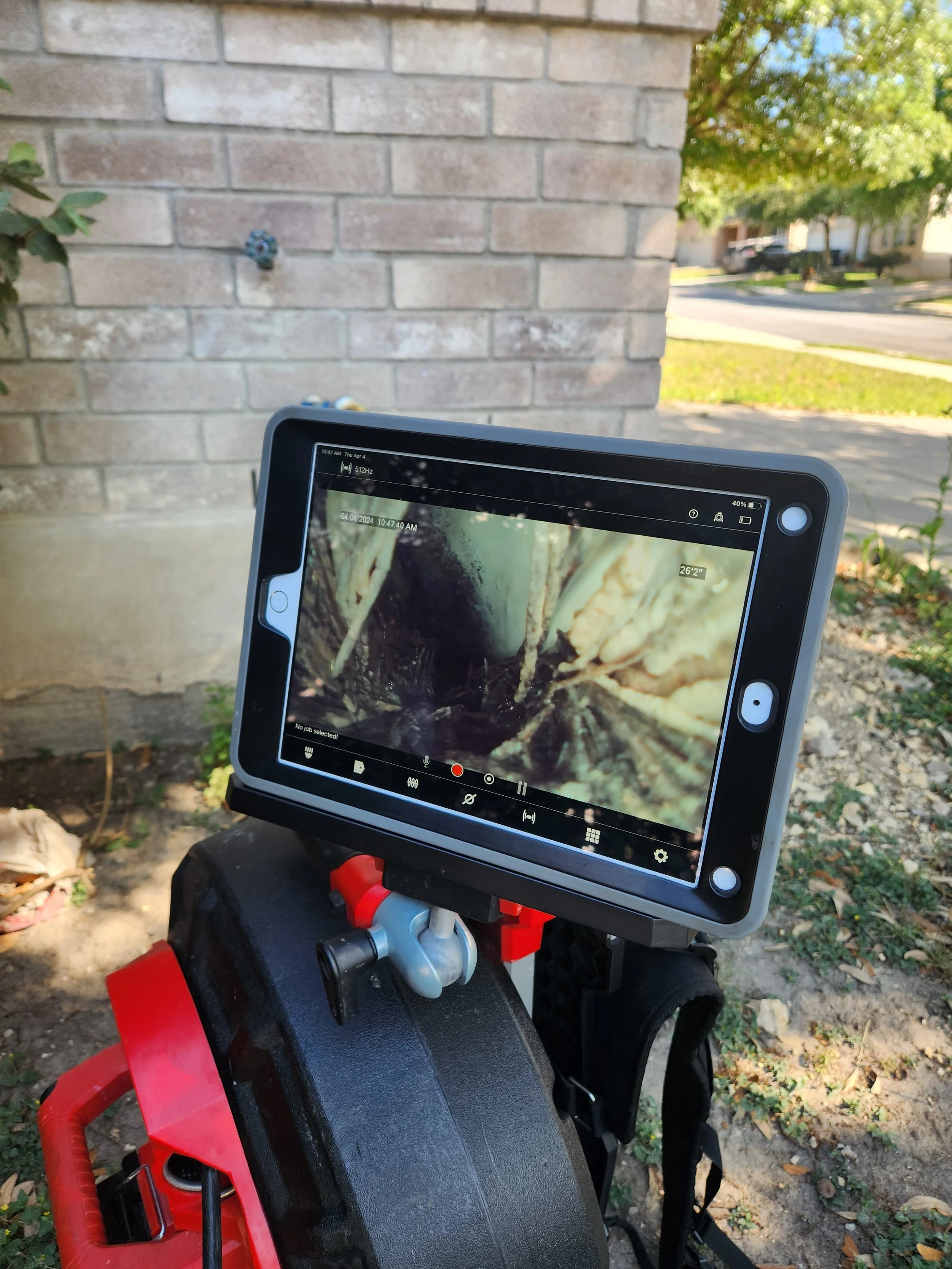 Close-up of a digital inspection device mounted on a work stand, displaying a camera view of a dark pipe, outdoors next to a brick wall with a sprinkler head, grass, trees, and a street in the background.