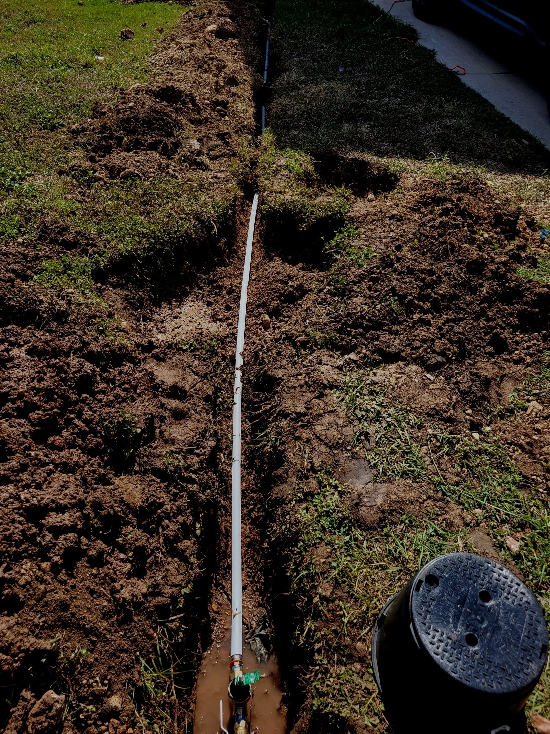 A partially excavated trench with a white pipe installed for plumbing or irrigation, with a black lid or cap nearby on the ground.