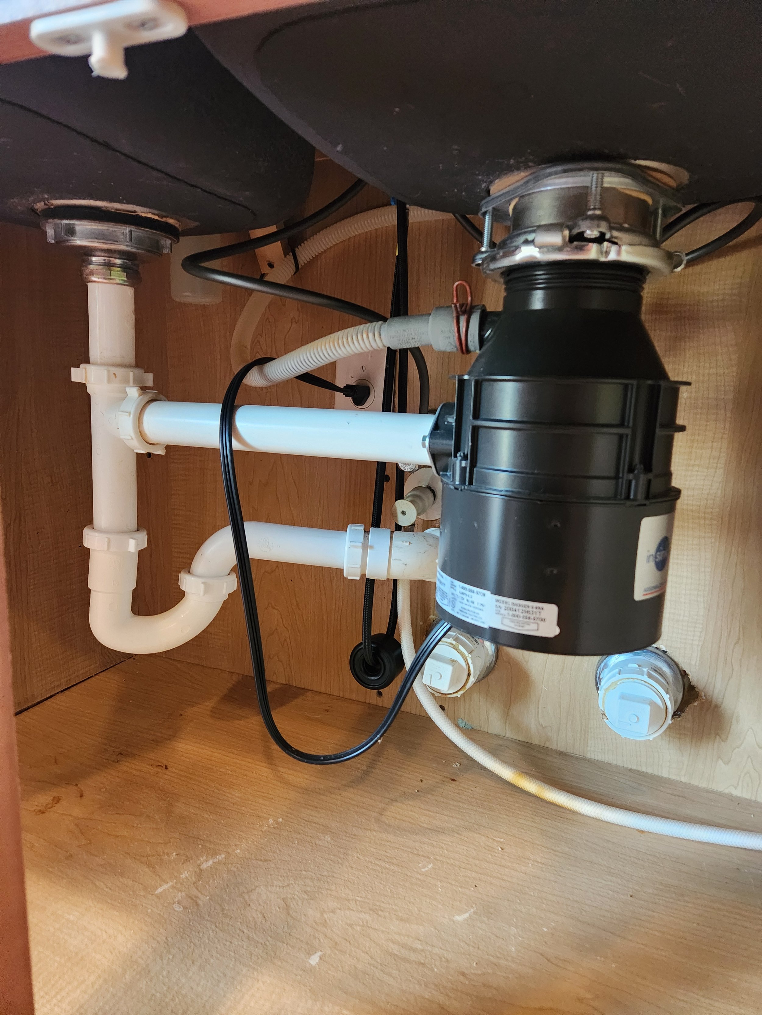 The image shows the plumbing system underneath a kitchen sink, including white PVC pipes, a black garbage disposal, and other fittings and hoses.