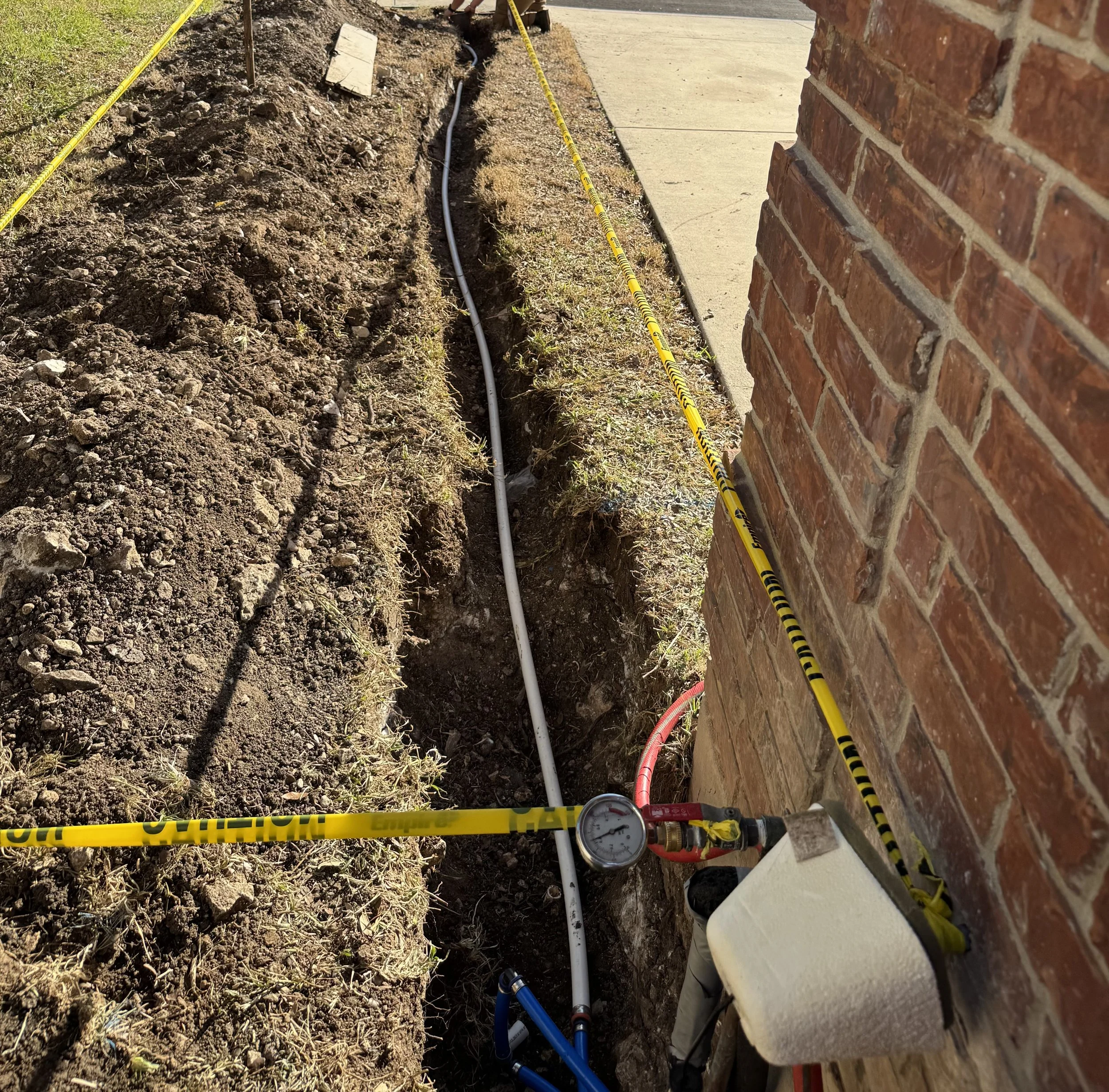 A partially excavated trench with a white pipe installed for plumbing or irrigation, with a black lid or cap nearby on the ground.