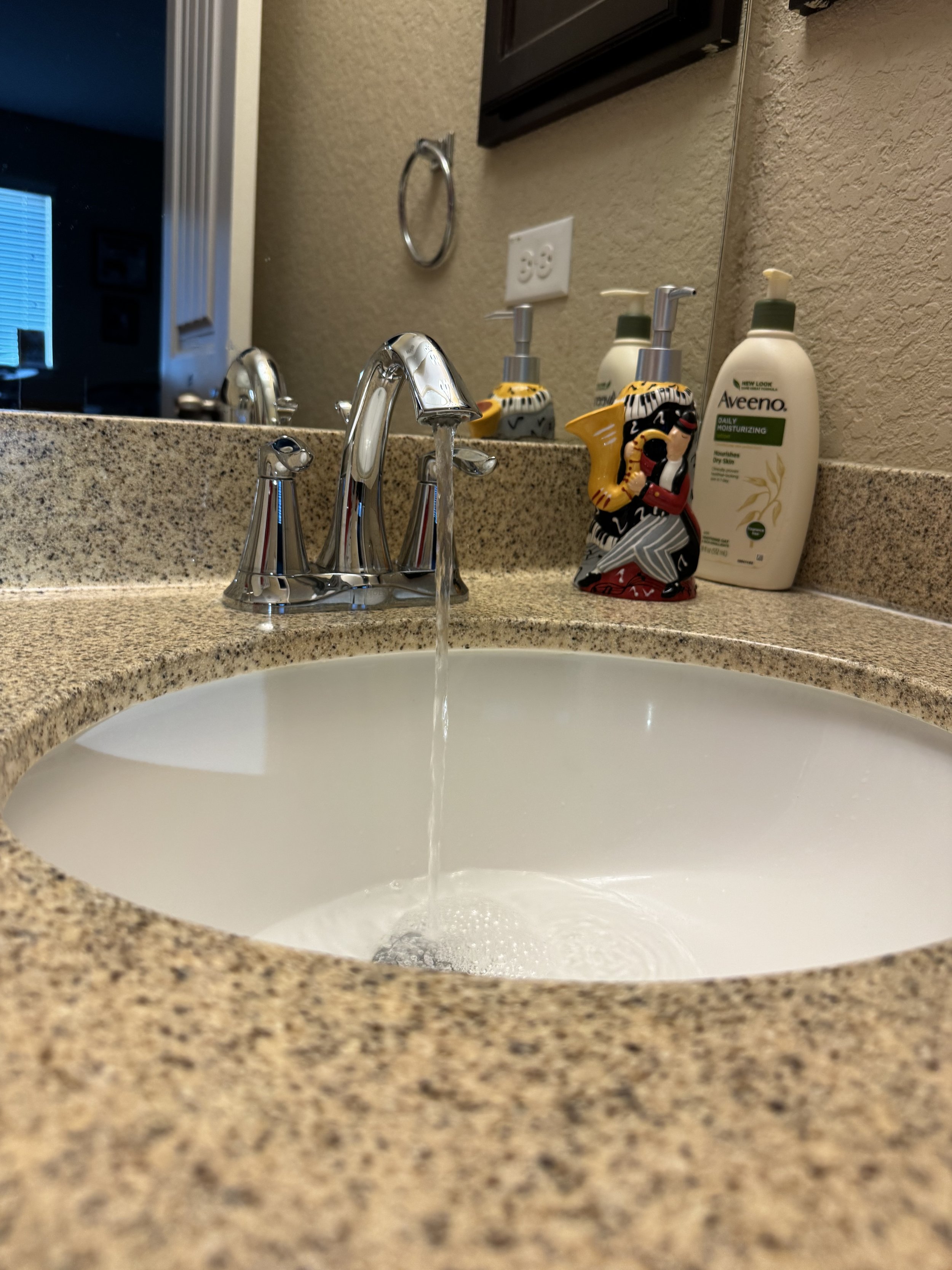 A bathroom countertop with a running faucet over a white oval sink, and various toiletries including a soap dispenser and a decorative figurine of a woman in colorful clothing.