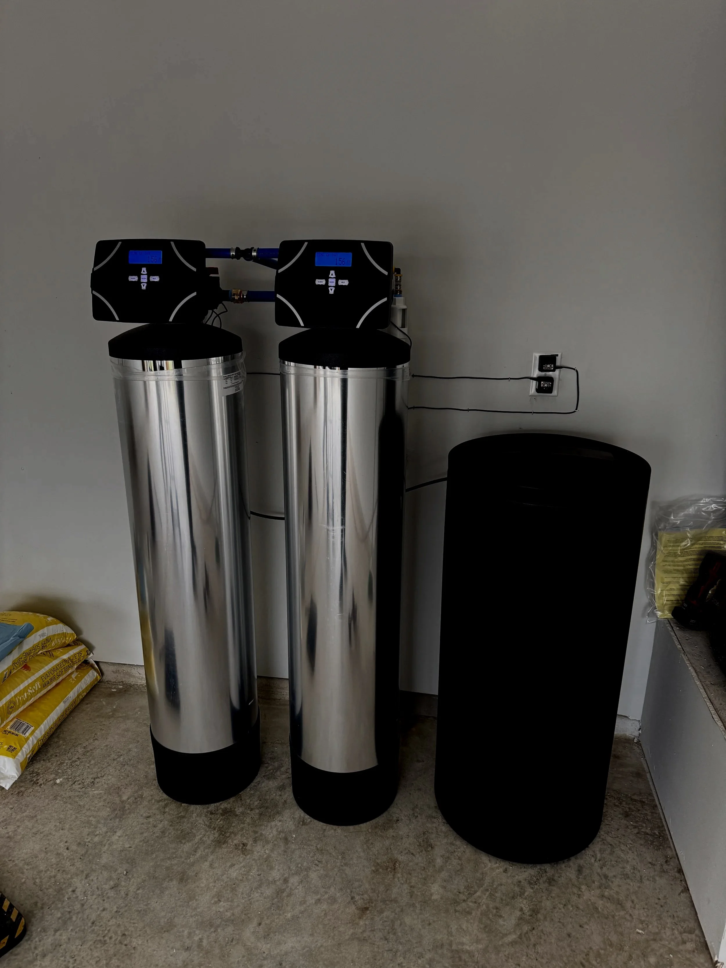 Water softening system with two tall stainless steel tanks and digital control heads installed on a white wall in a utility room.
