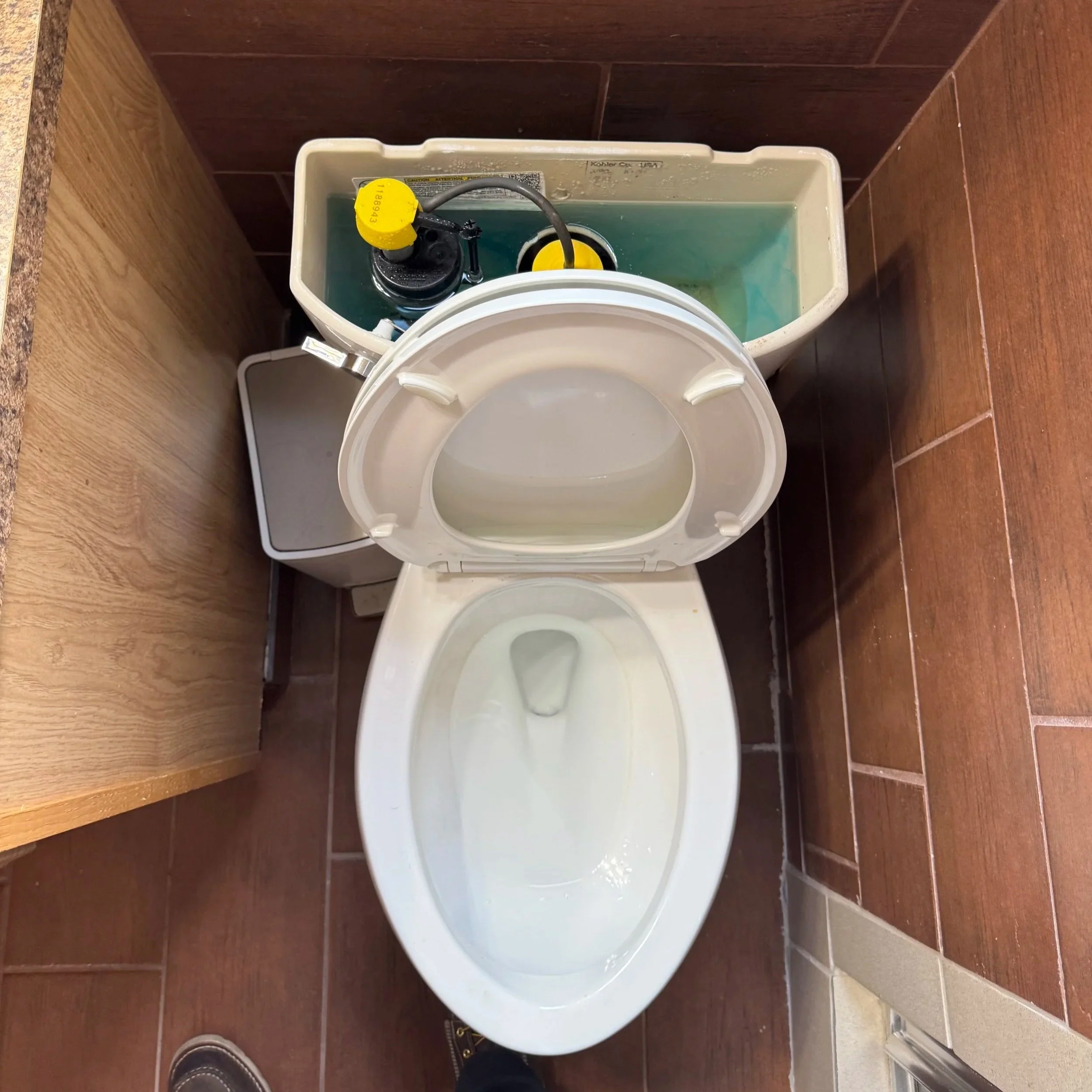 A toilet with the lid open, small trash bin to the side, and a tank with a water flushing mechanism behind it. The floor is tiled in brown and the walls have brown tiles.
