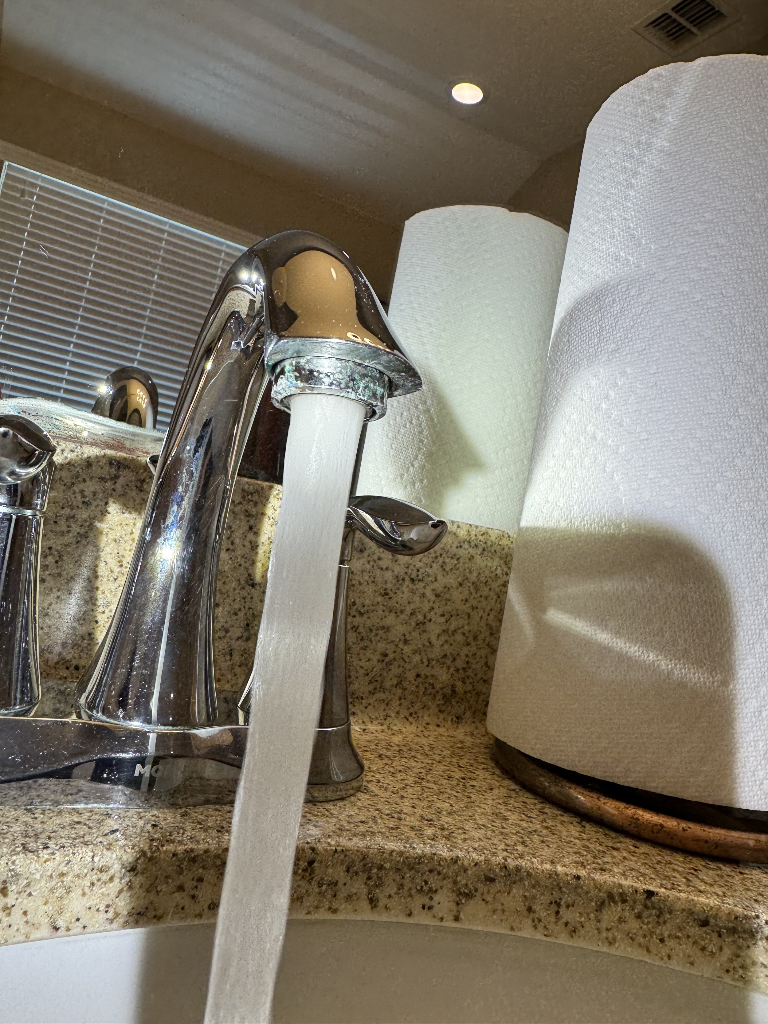 Running water flows from a kitchen faucet onto a granite countertop, with paper towels and a paper towel holder nearby.
