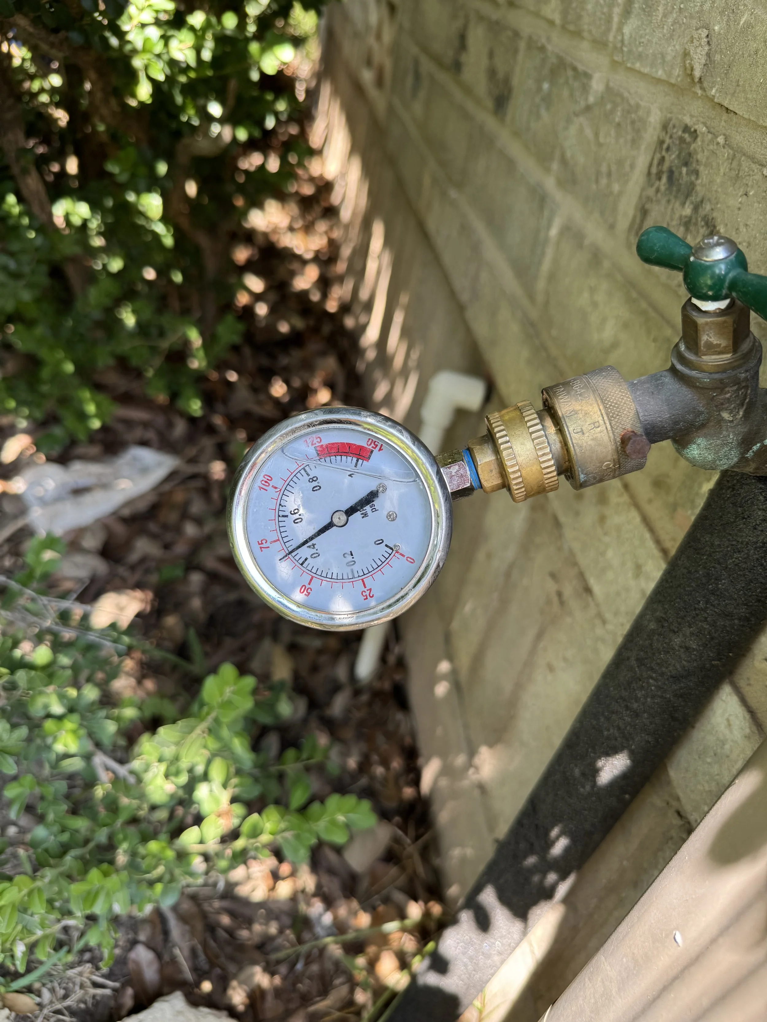 A pressure gauge attached to a water faucet next to a brick wall and some green plants.