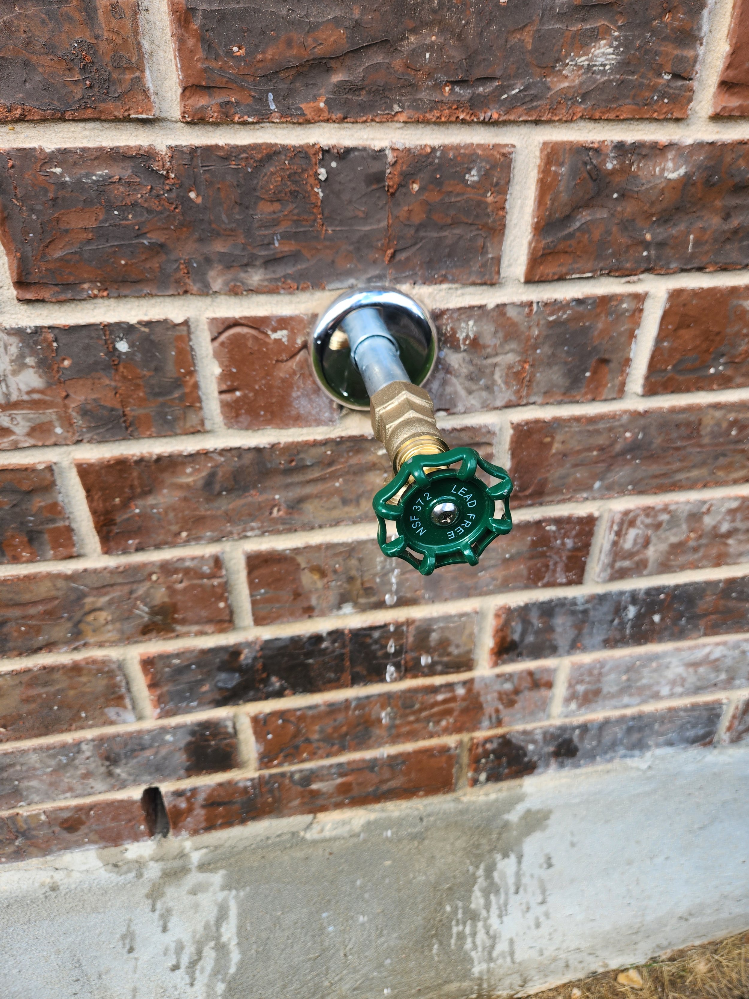 A water spigot with a green handle attached to a brick wall, with water dripping from the spout.