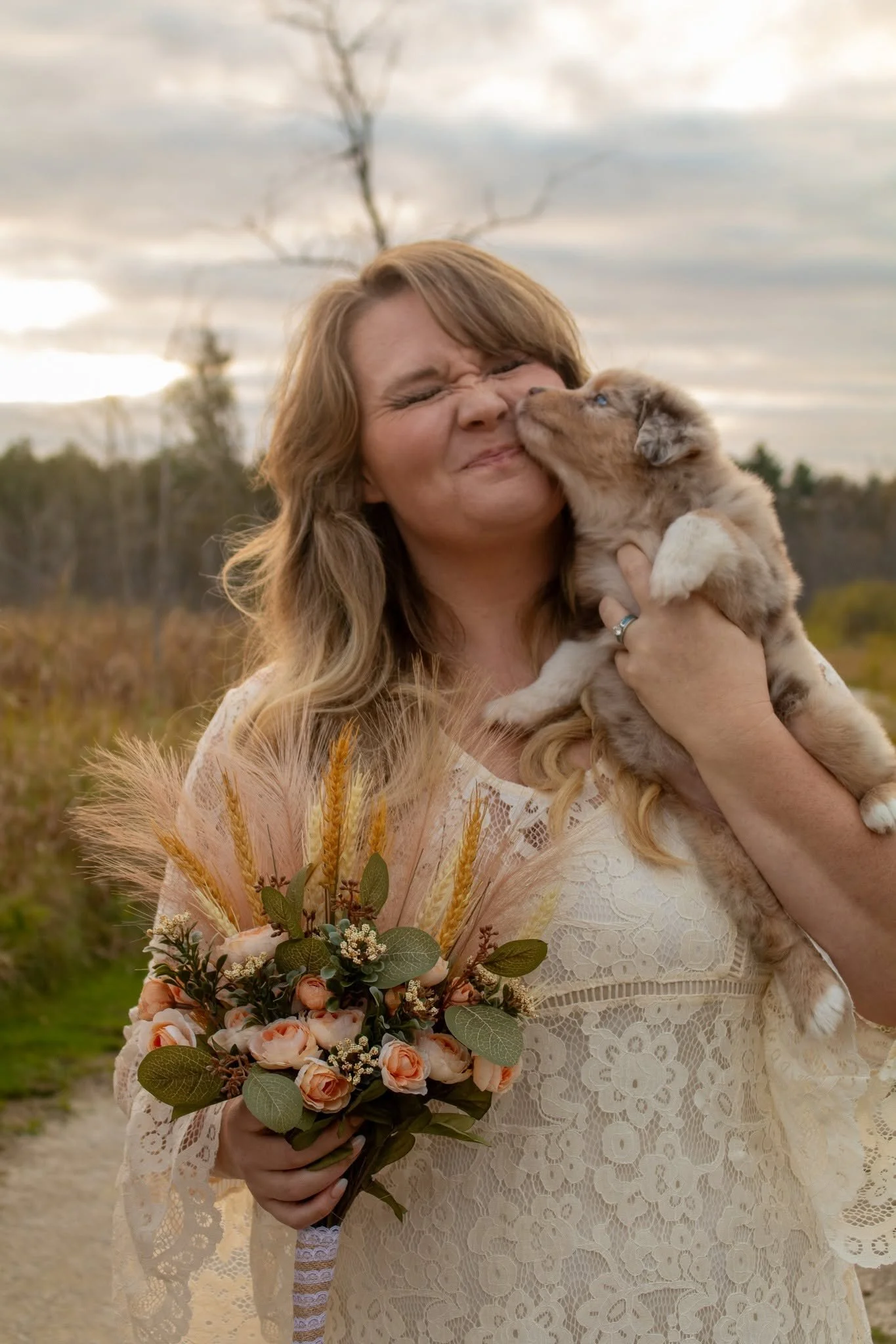 A woman with long blonde hair in a lace dress holding a bouquet of flowers, standing outdoors in a field with a cloudy sky and leafless trees in the background. She is smiling and kissing a puppy.