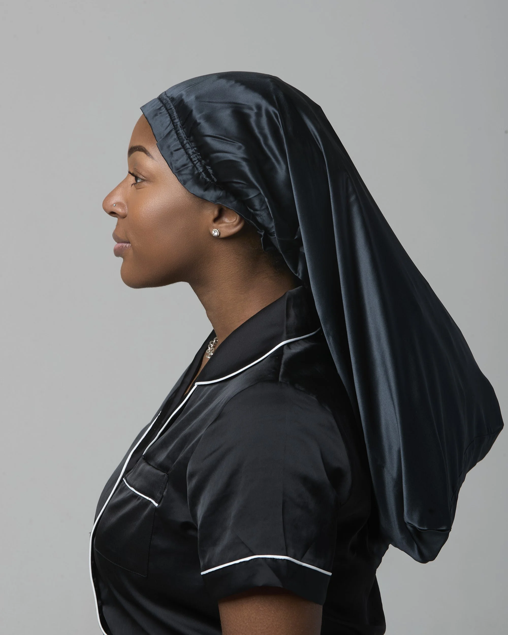 Side profile of a woman wearing a shiny black satin bonnet, with a nose piercing and diamond earring, against a plain gray background.