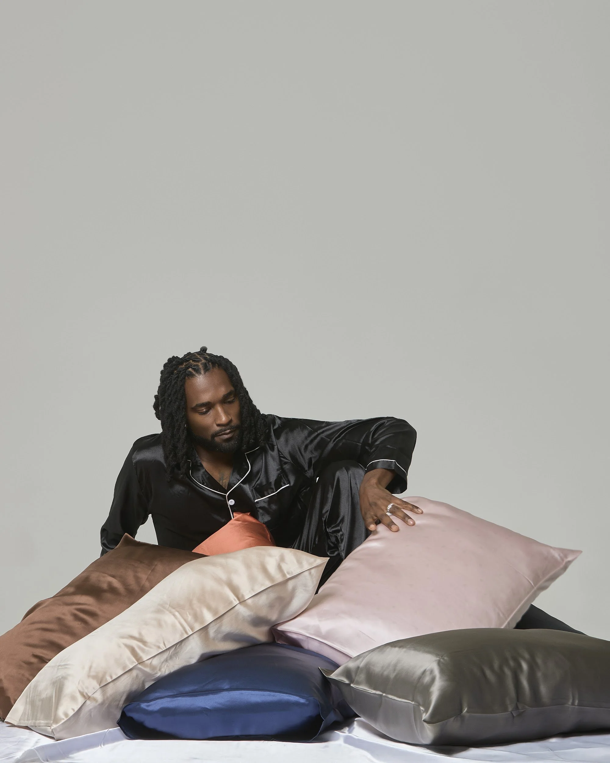 A man with long dreadlocks and facial hair, wearing a black satin shirt, is sitting on a bed surrounded by pillows of various colors including brown, beige, blue, pink, and gray, against a plain light gray background.