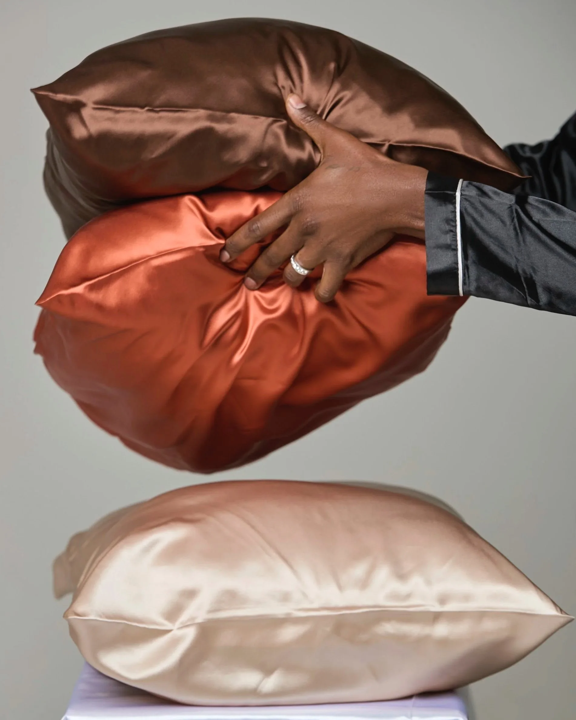Three 100% mulberry silk pillows in shades of coffee brown, rust/burnt orange, and beige stacked on each other against a grey background.