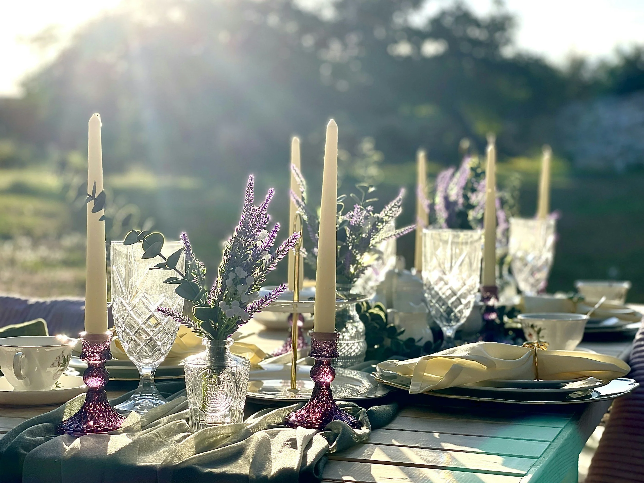 A table setting outdoors with candles, crystal glasses, floral arrangements, plates, and napkins, illuminated by sunlight.