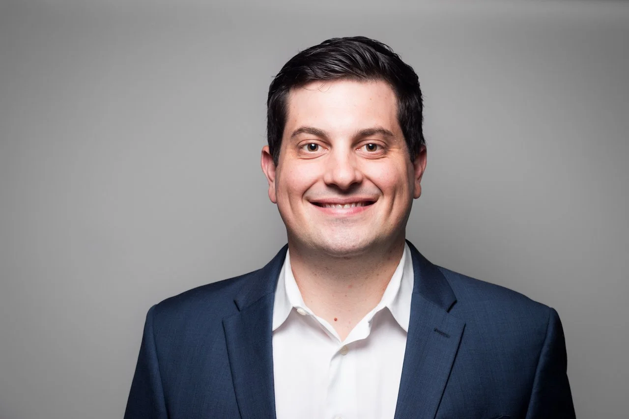 Professional headshot of a smiling man in a navy suit and white shirt against a gray background.