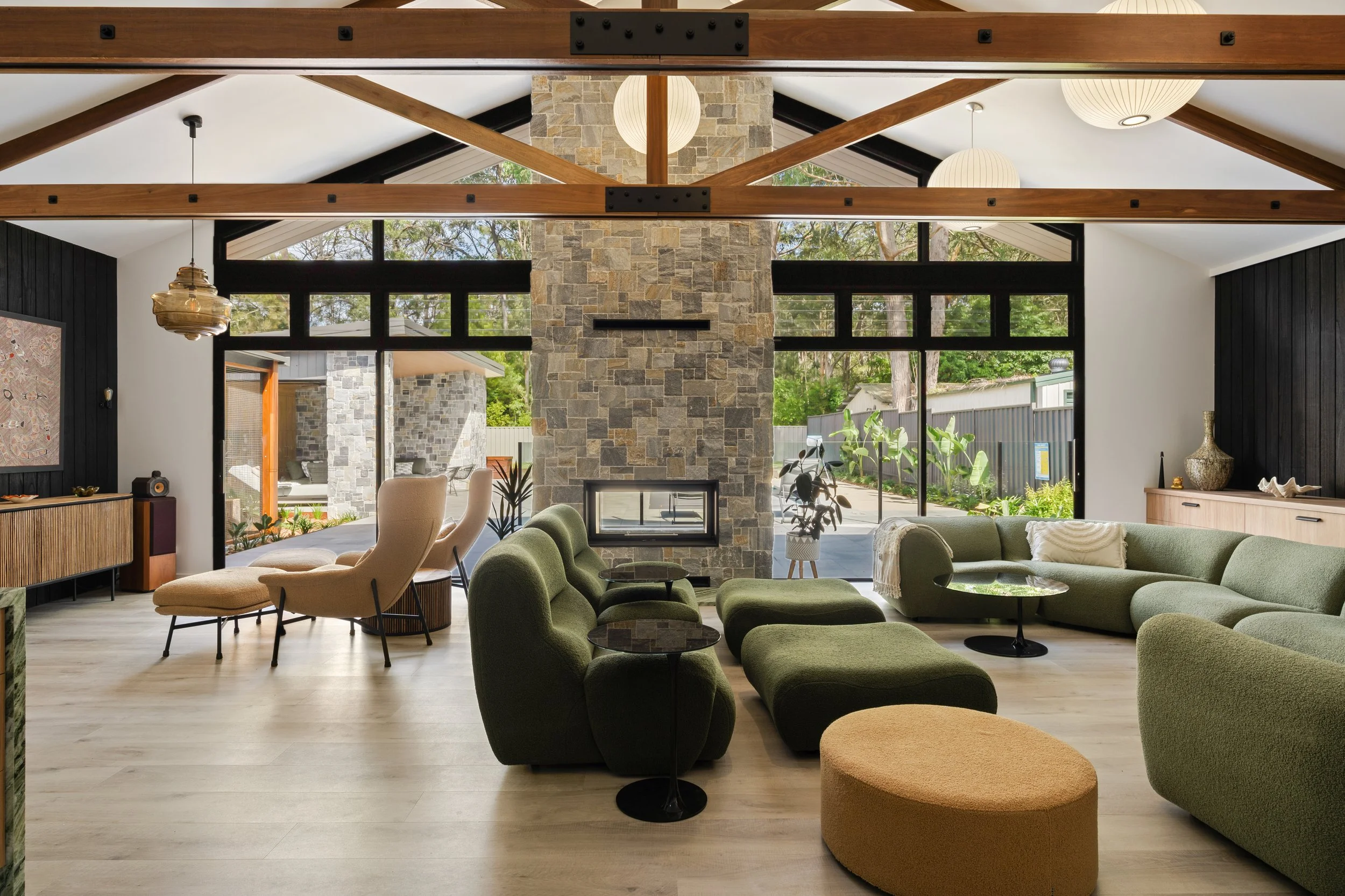Living room with large windows, stone fireplace, green sectional sofa, beige armchairs, and wooden beams.
