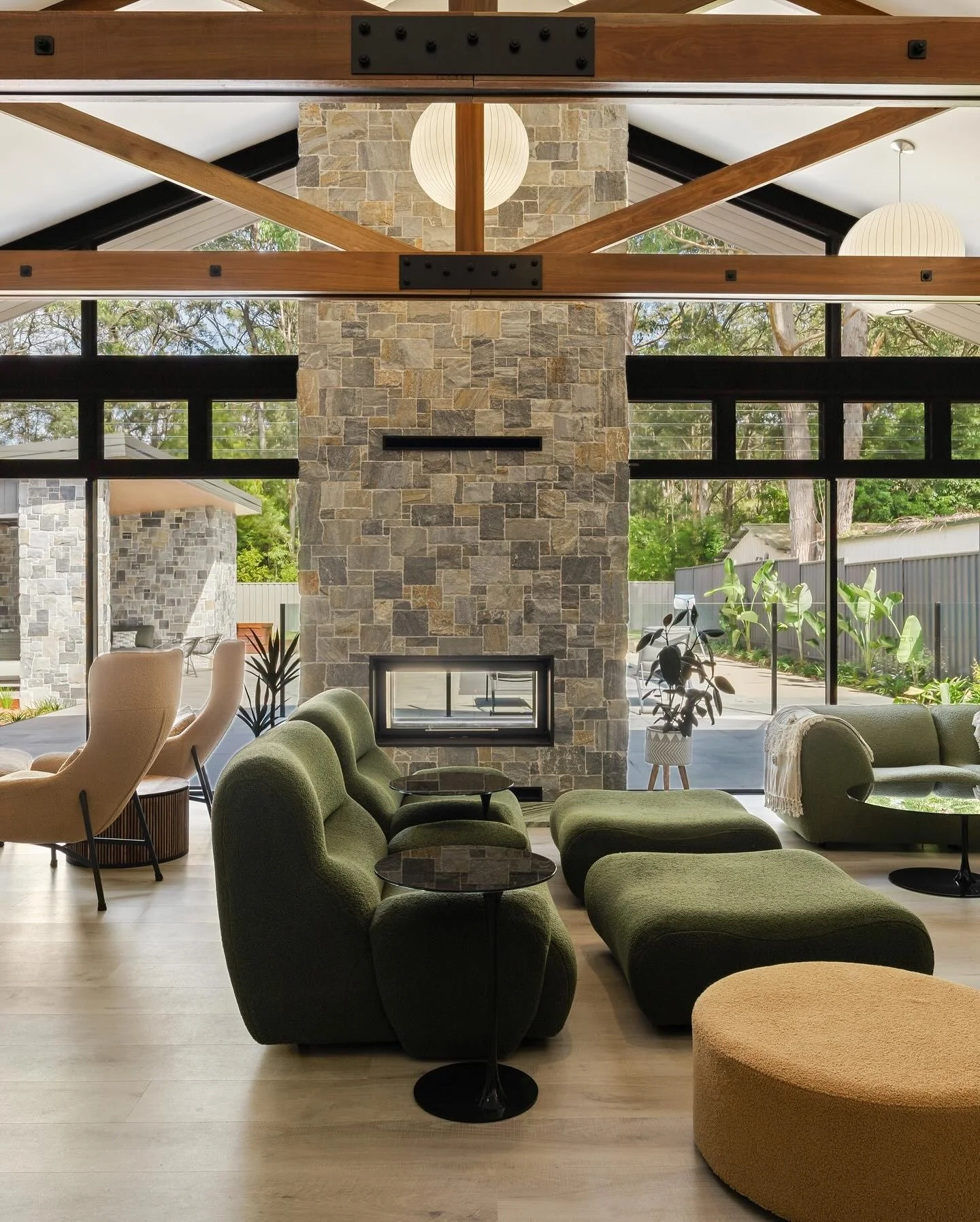 Modern living room with green and beige armchairs, a stone fireplace, floor-to-ceiling windows, and indoor plants, with a view of the outdoor patio and garden.
