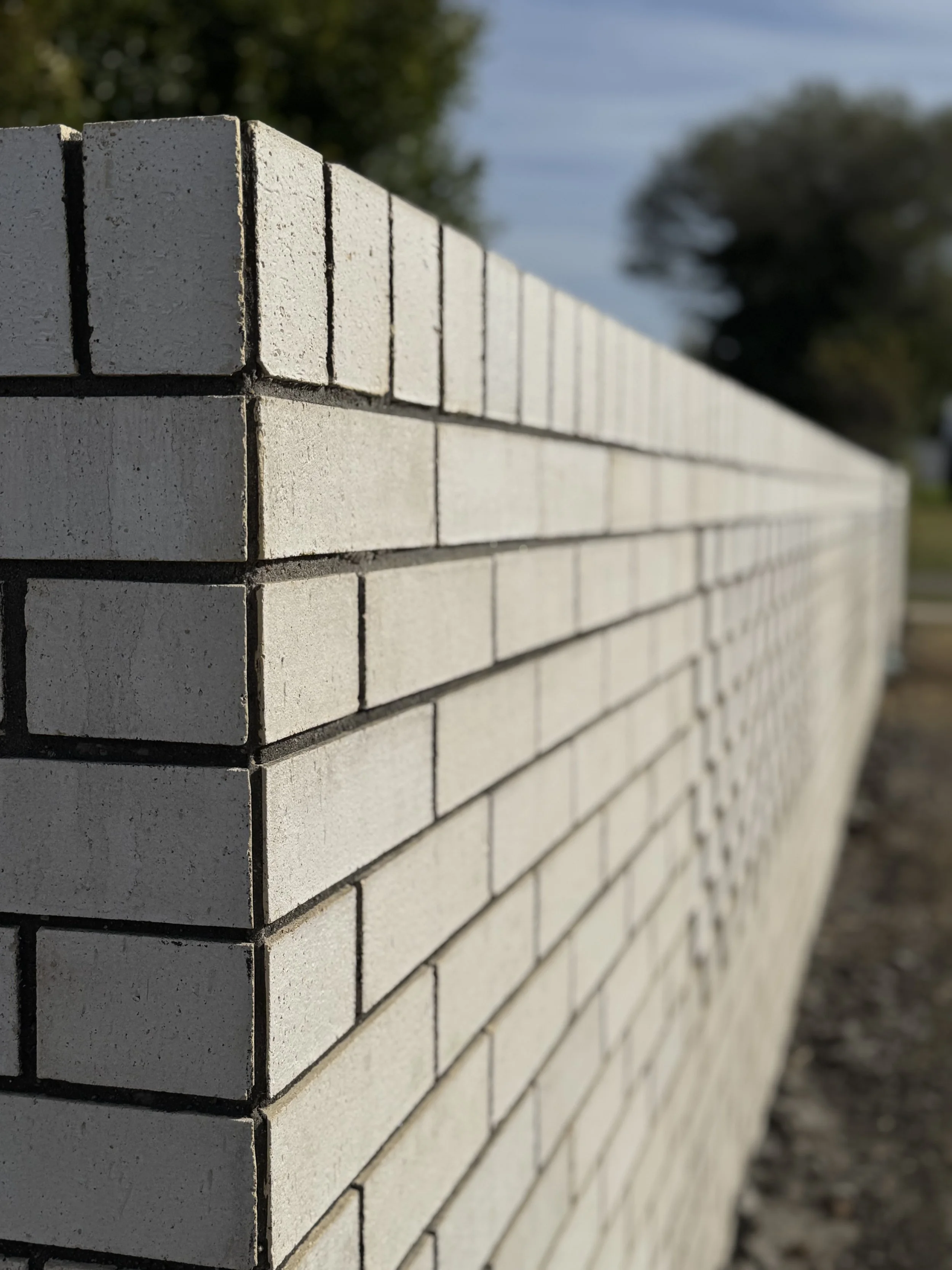 Close-up view of a white brick wall with black mortar lines, stretching into the distance, with trees and sky in the background.