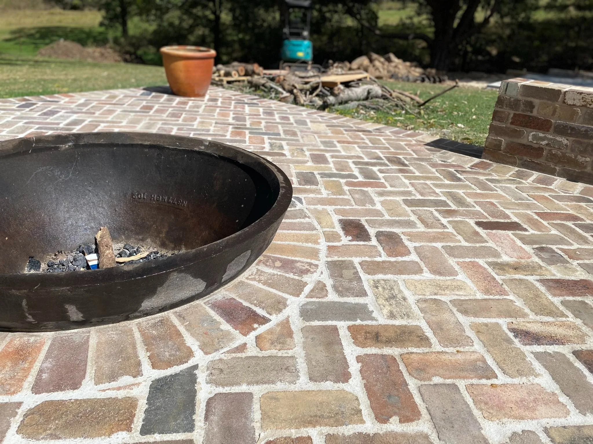 Brick outdoor patio with a black fire pit containing ashes, a terracotta pot in the background, and a grassy yard with trees.
