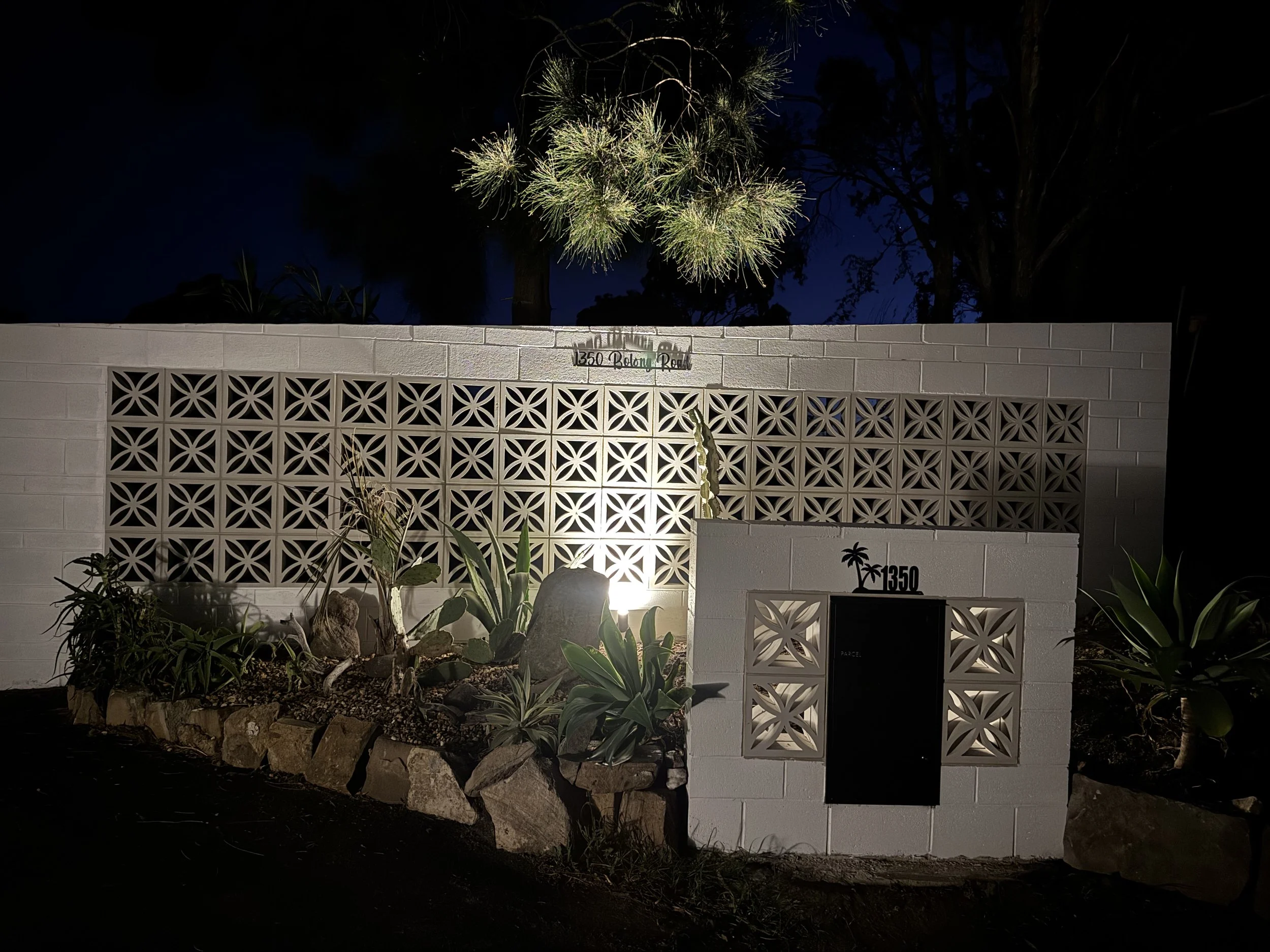 Nighttime view of a white brick wall with decorative blocks, a black mailbox, and the street number 1350. In front, there is a garden with rocks and various succulents. A tree is visible in the background, illuminated by a light.
