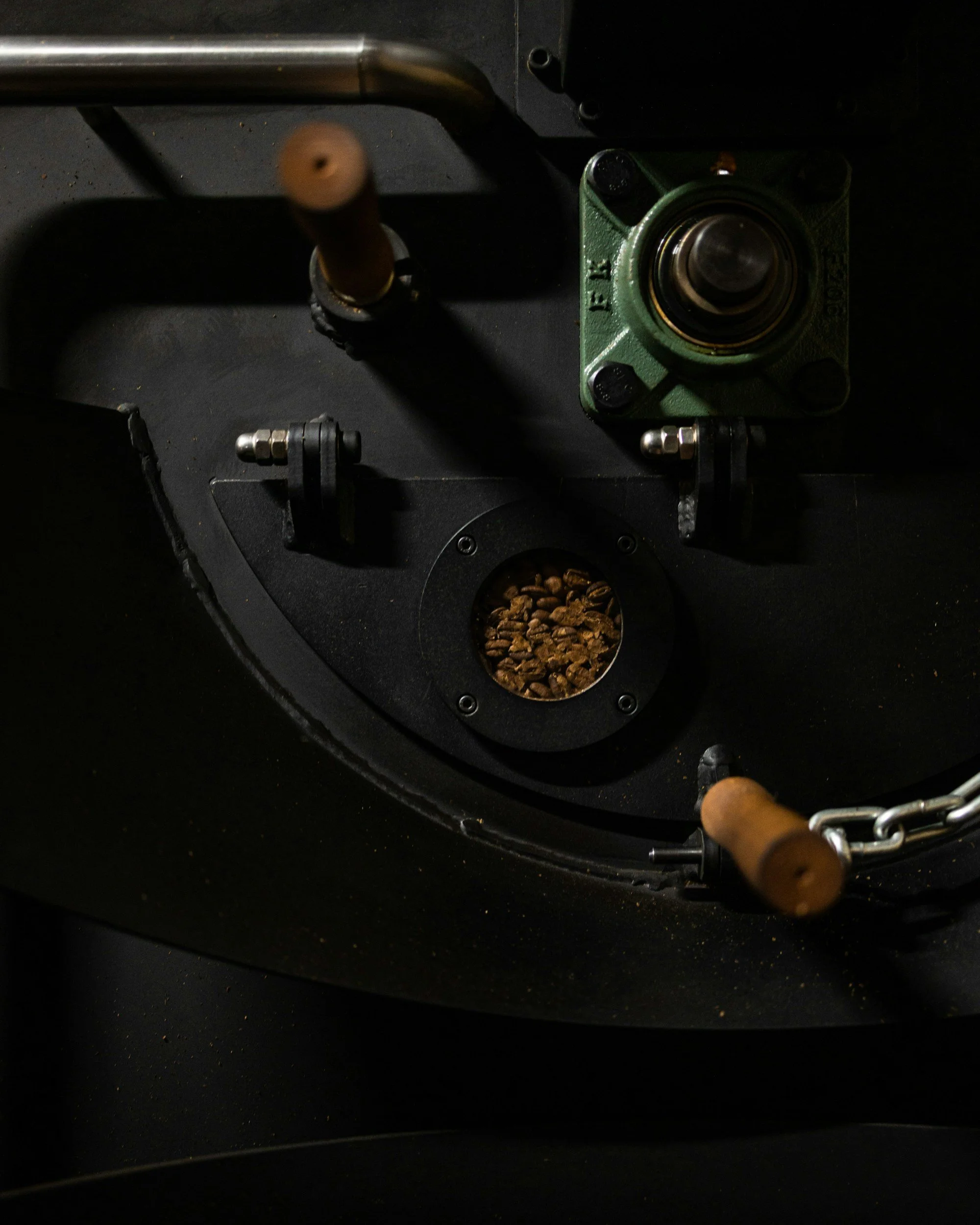 Close-up of a coffee roasting machine, showing roasted coffee beans inside the roaster, with metal and wooden handles and parts of the machinery visible.