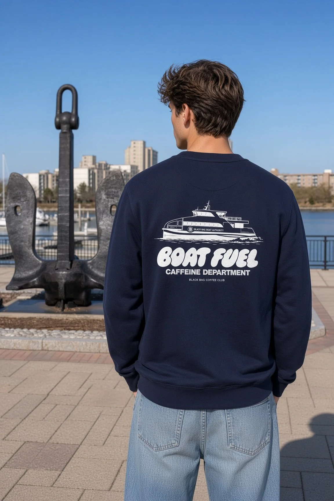Back of a person wearing a navy sweatshirt with a boat illustration and text related to boat fuel and caffeine department, standing near a large black anchor sculpture by a waterfront with city buildings in the background.