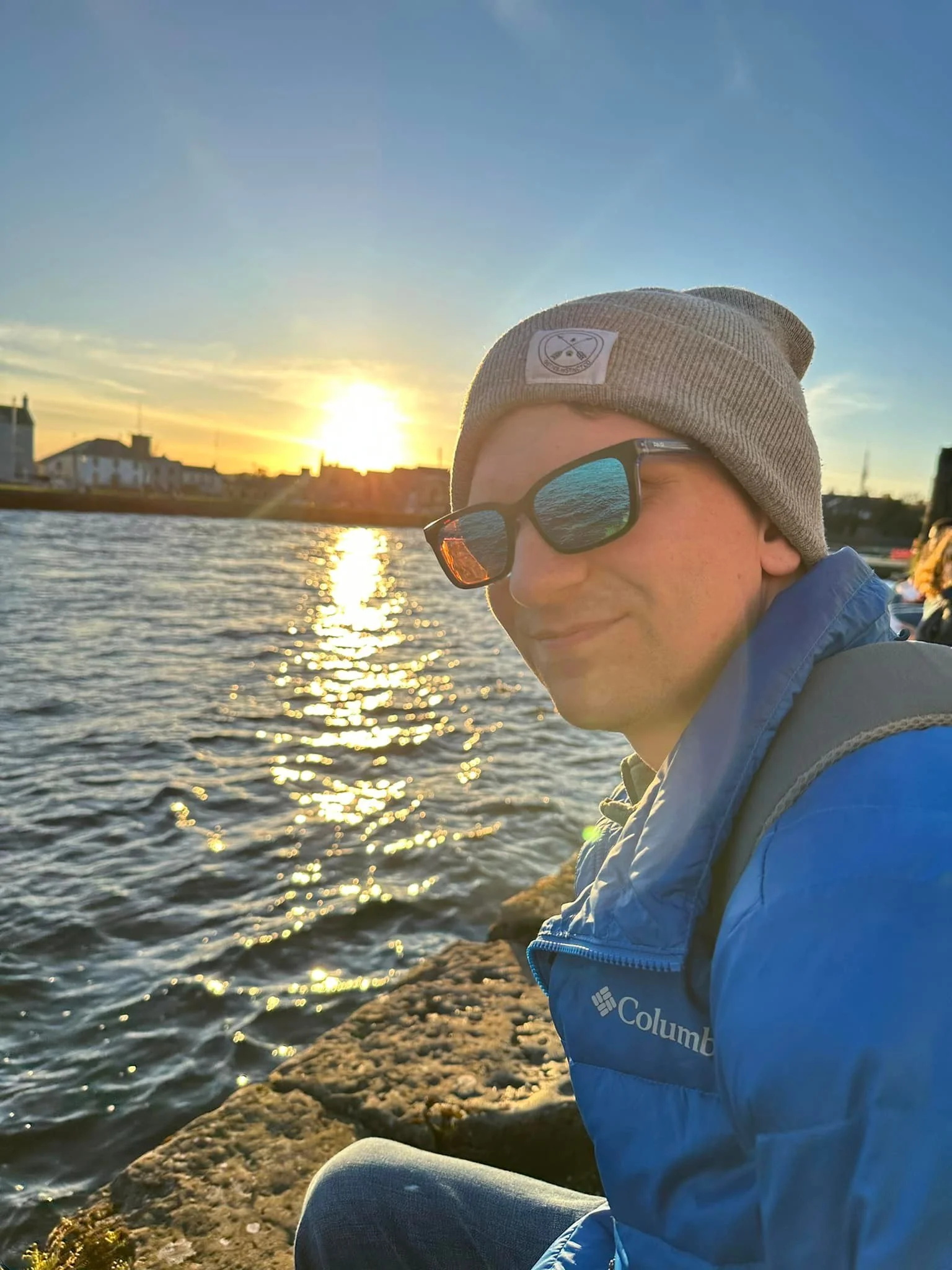 A young man wearing sunglasses and a gray beanie hat sitting by the water during sunset, with a slight smile on his face.