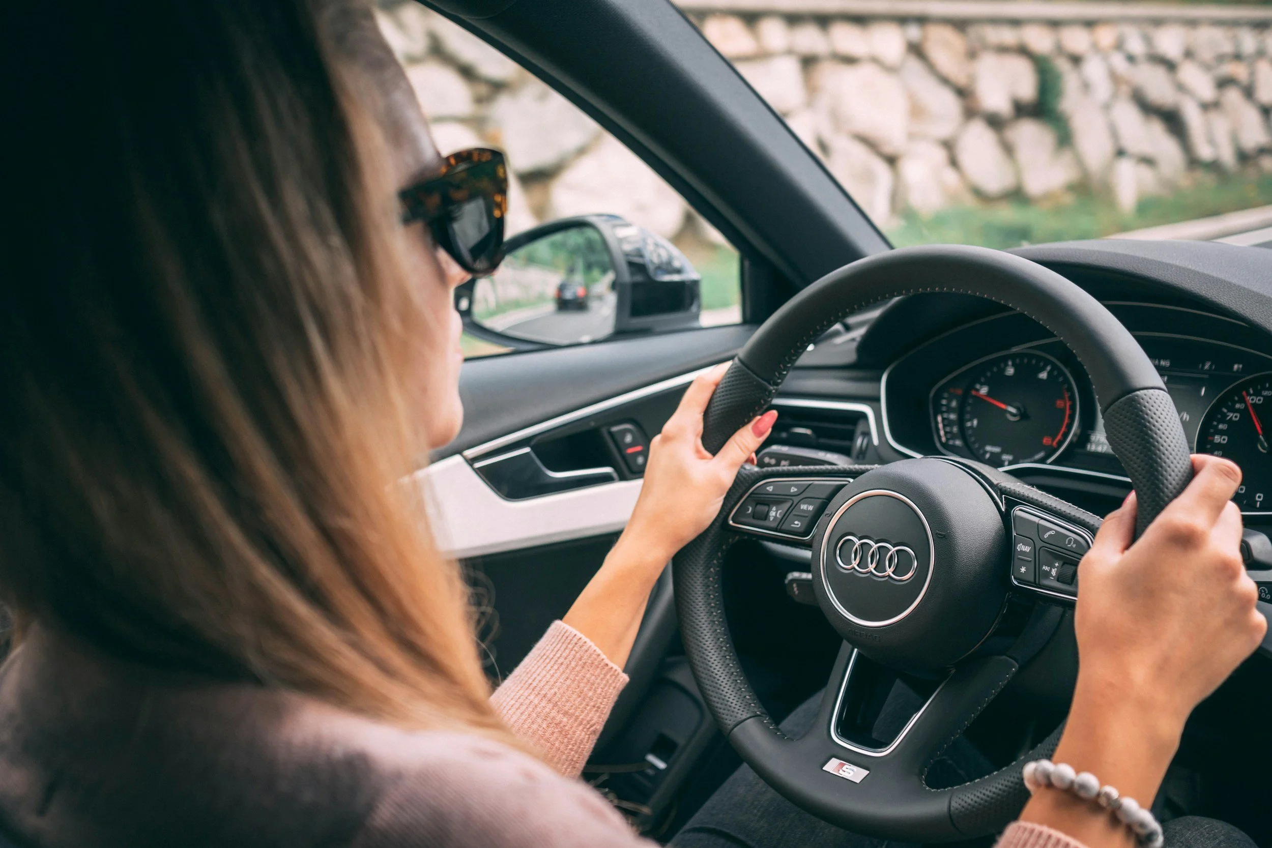 A woman driving an Audi vehicle, wearing sunglasses, with her left hand on the steering wheel and her right hand on the gear shift. She is on a road with a stone wall on the side.