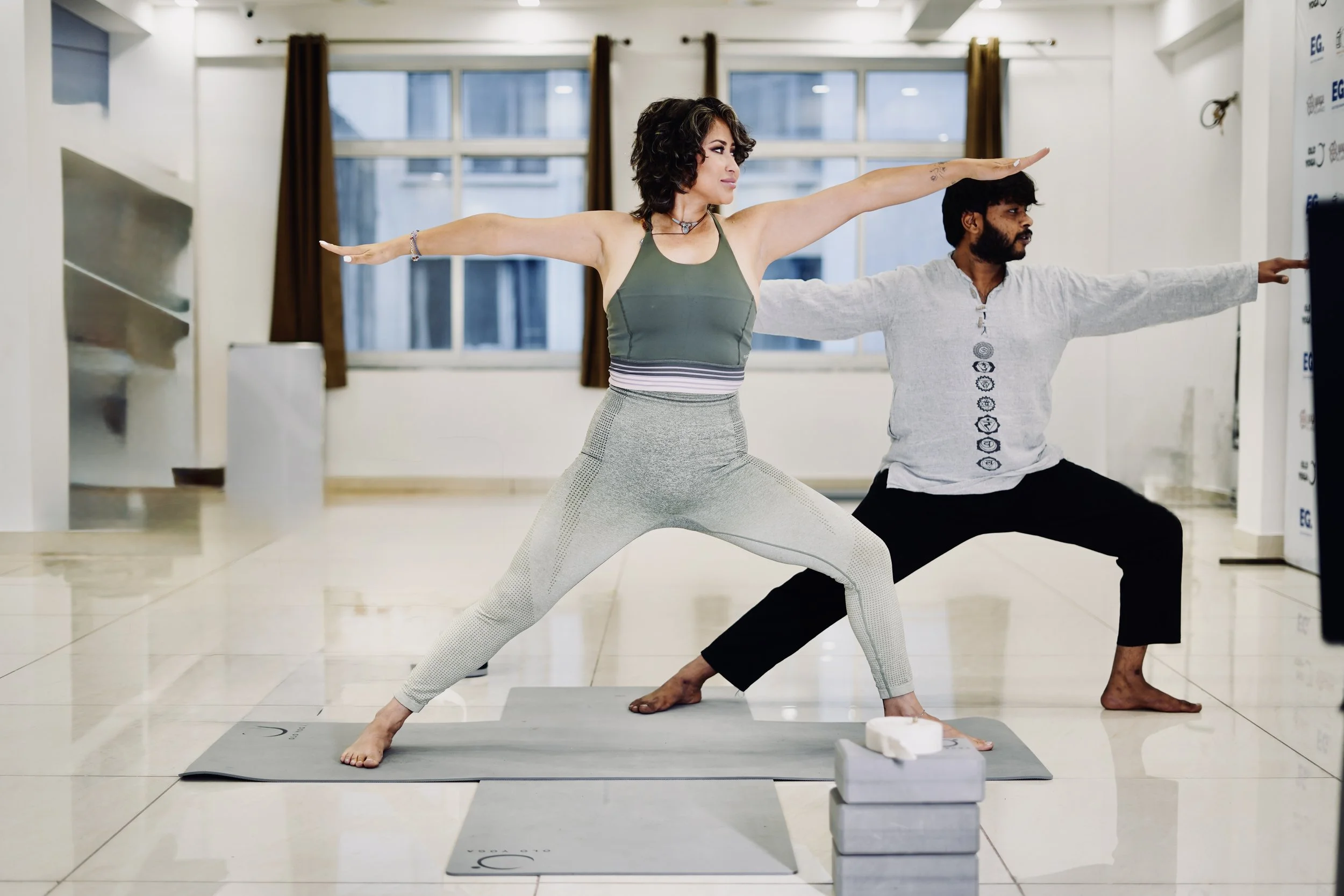 Woman and man practicing yoga indoors; woman in Warrior 2 pose, man in modified Warrior pose, both on yoga mats and smiling.