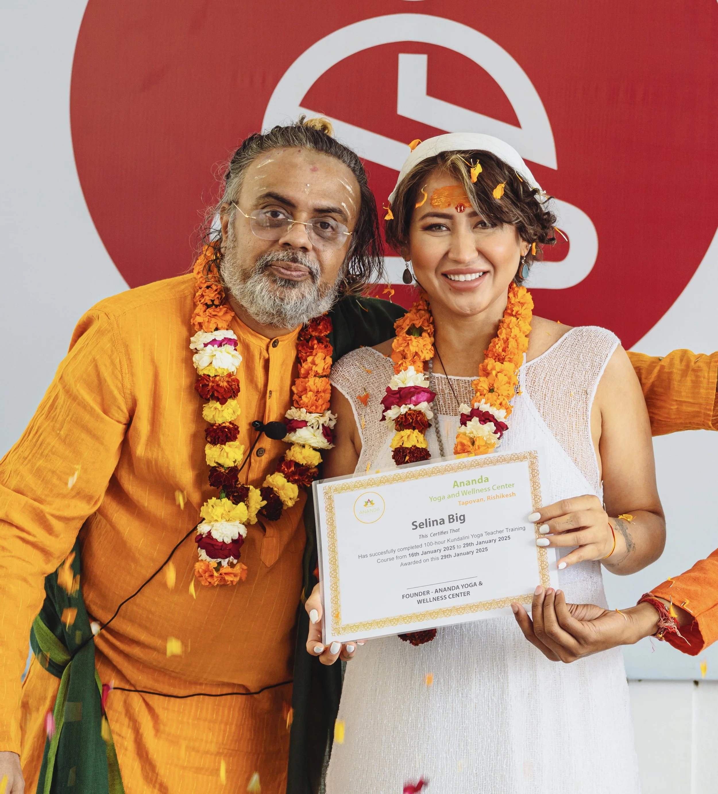 Yoga instructor receiving her Teacher Training certificate and smiling, standing next to guru in traditional Indian attire. They are wearing flower necklaces and garlands. The woman has flower petals in her hair and traditional jewellery.