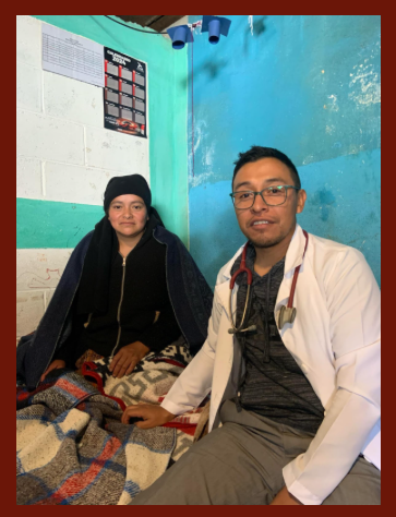 A doctor wearing a white coat and stethoscope sitting next to a woman, who is wrapped in a blanket, in a medical setting with blue walls.