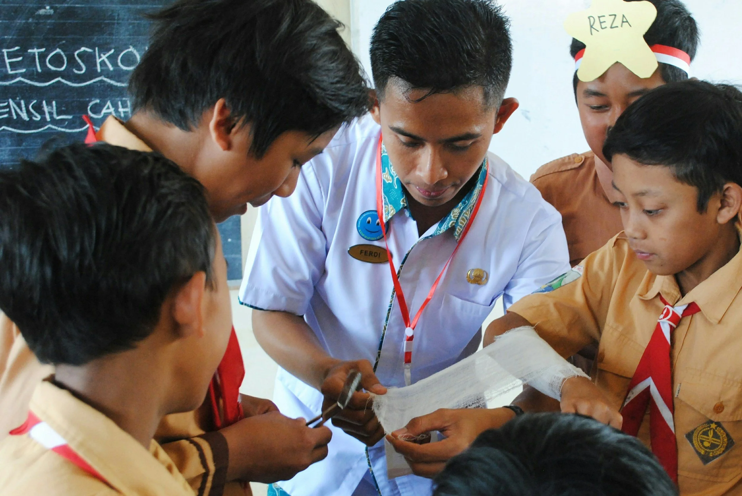 A teacher and several students, all wearing scouts uniforms, are gathered closely together in a classroom. The teacher, wearing a white shirt with a name tag, is showing something on a piece of cloth to the students, who are attentively watching.